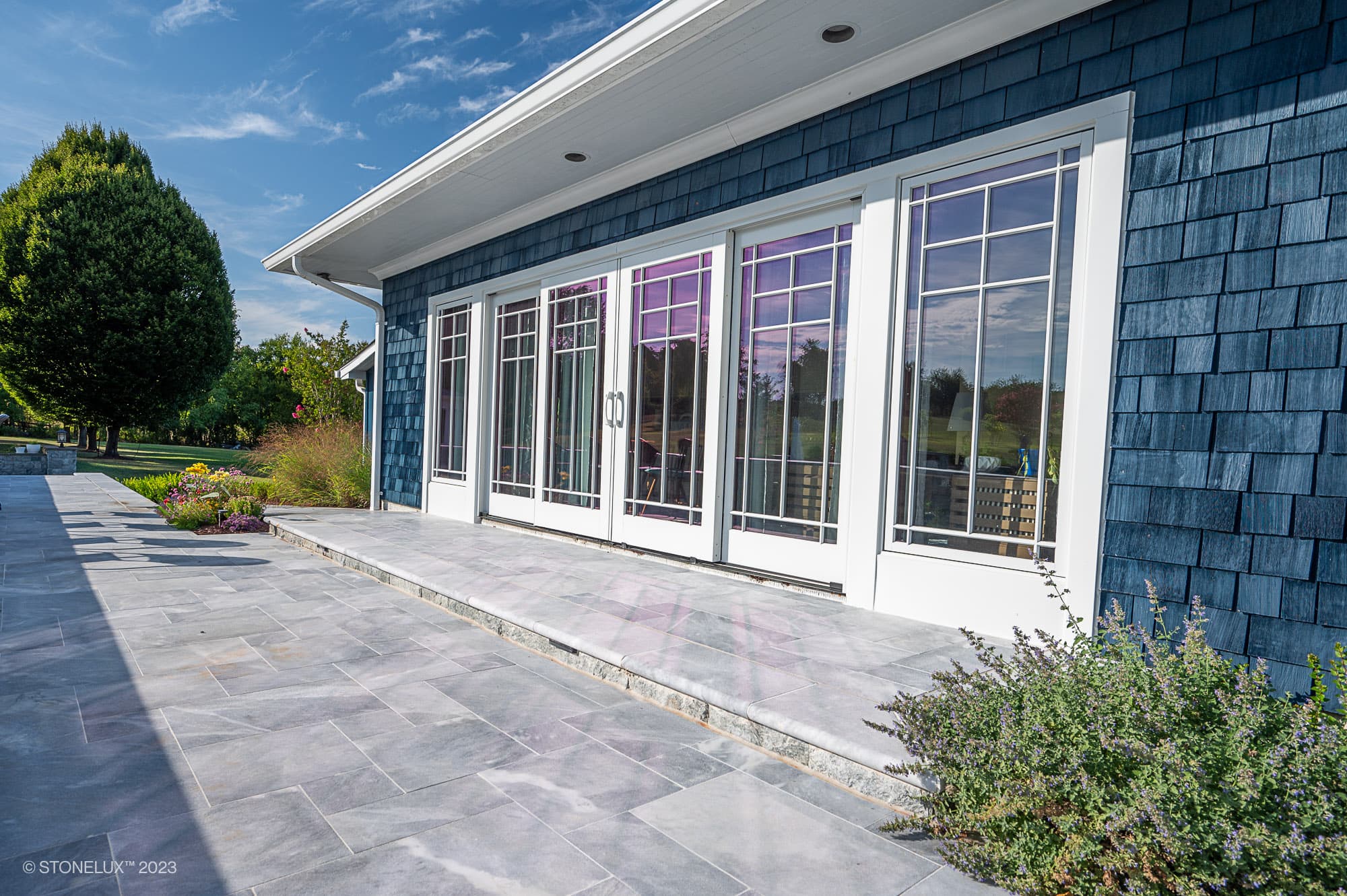 A modern house with blue shingle siding and large glass doors opens to a spacious stone patio, featuring the Blue Haven: The Grey and Blue Sandblasted Marble Pool Project, bordered by plants and trees under a partly cloudy sky.