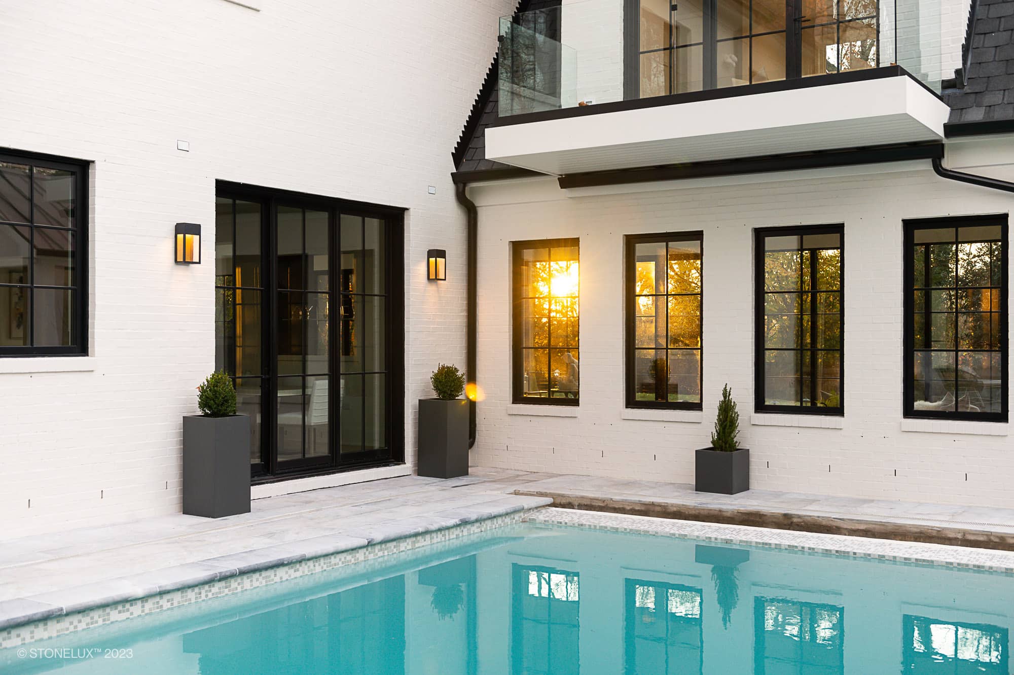 Modern house exterior with large black-framed windows, white brick walls, and small potted plants. The Arctic Elegance: Blue and Grey Sandblasted Marble Pool and Patio Project gleams in the foreground, reflecting the golden sunset.