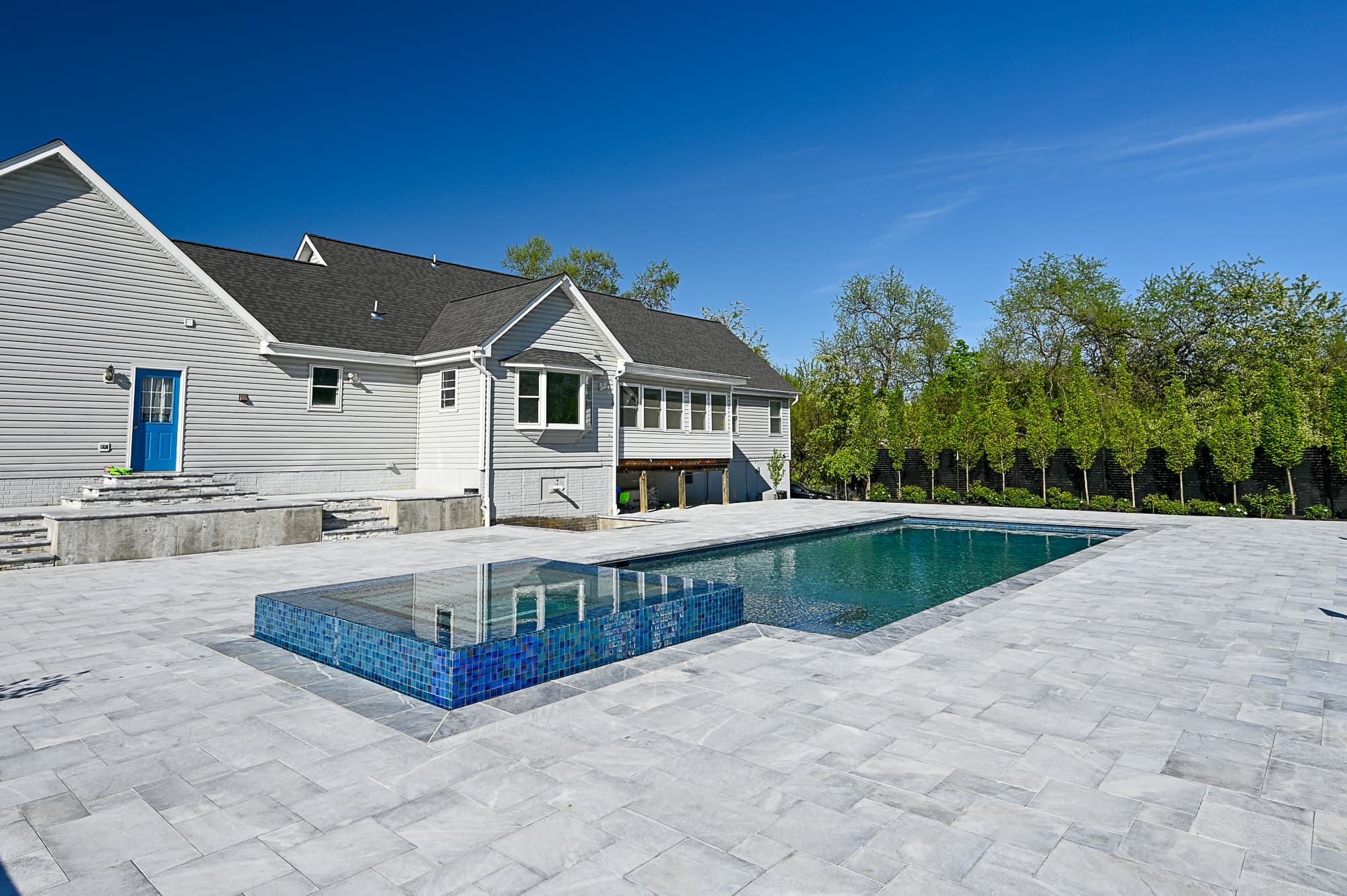 A modern backyard, inspired by Arctic Retreat, features a rectangular swimming pool and raised spa, surrounded by luxury Arctic Grey Marble pavers. The light-colored house with a blue door overlooks trees lining the yard’s edge.