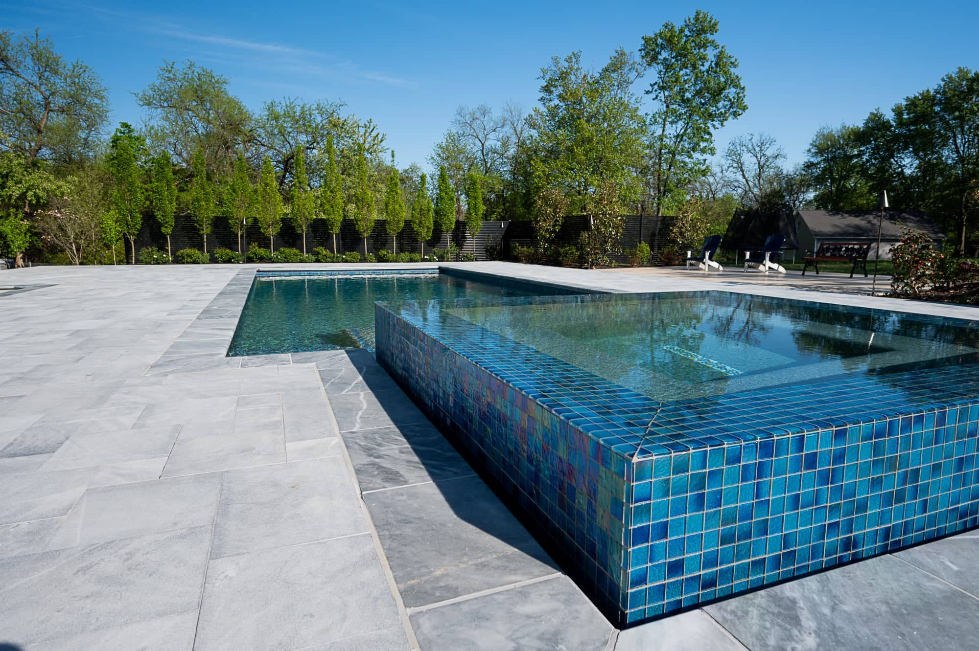 A modern outdoor swimming pool with a raised, blue-tiled square hot tub is surrounded by Arctic Retreat: Luxury Pool and Patio with Arctic Grey Marble Pavers and lush green trees under a clear blue sky.