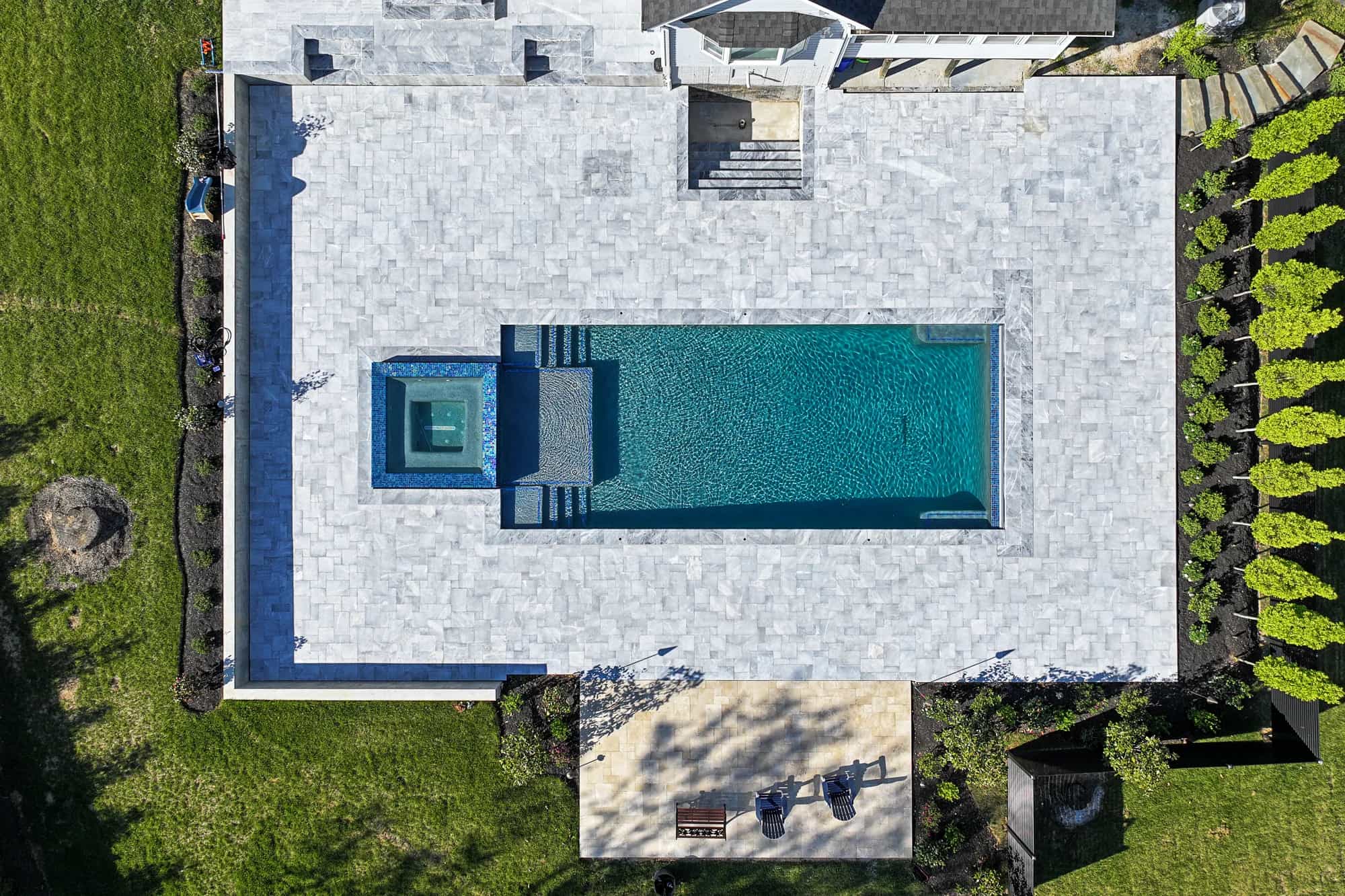 Aerial view of an Arctic Retreat: luxury pool and patio with Arctic Grey Marble Pavers, featuring a rectangular backyard swimming pool, attached square hot tub, lush grass, pristine landscaping, two lounge chairs, and a small table nearby.