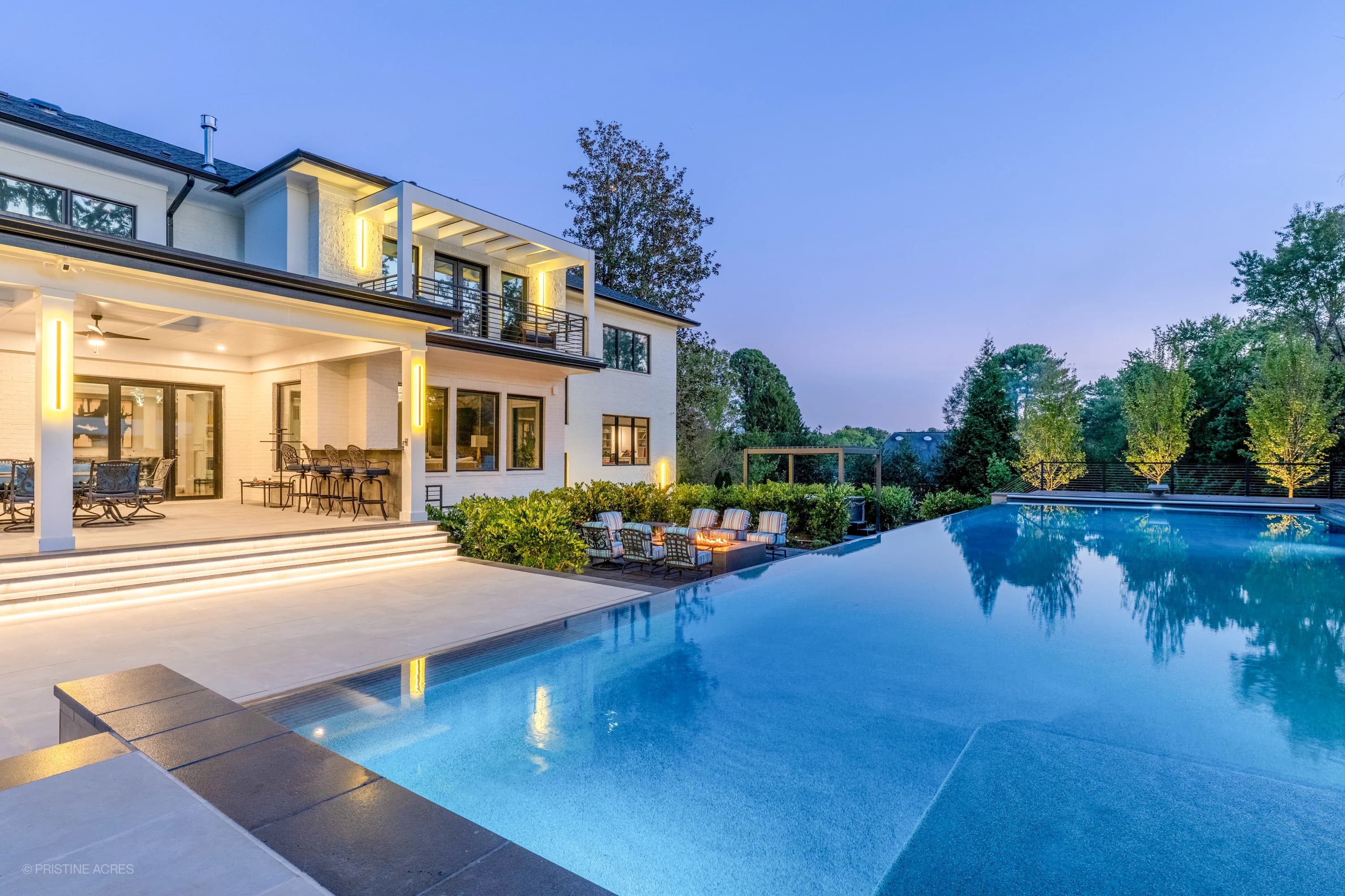 Modern McLean Pool Project in McLean VA features Black Basalt coping and Nexus White Porcelain pavers. The contemporary design includes a serene blue pool, lush landscaping, outdoor seating, and ambient lighting, creating a luxurious backyard oasis.