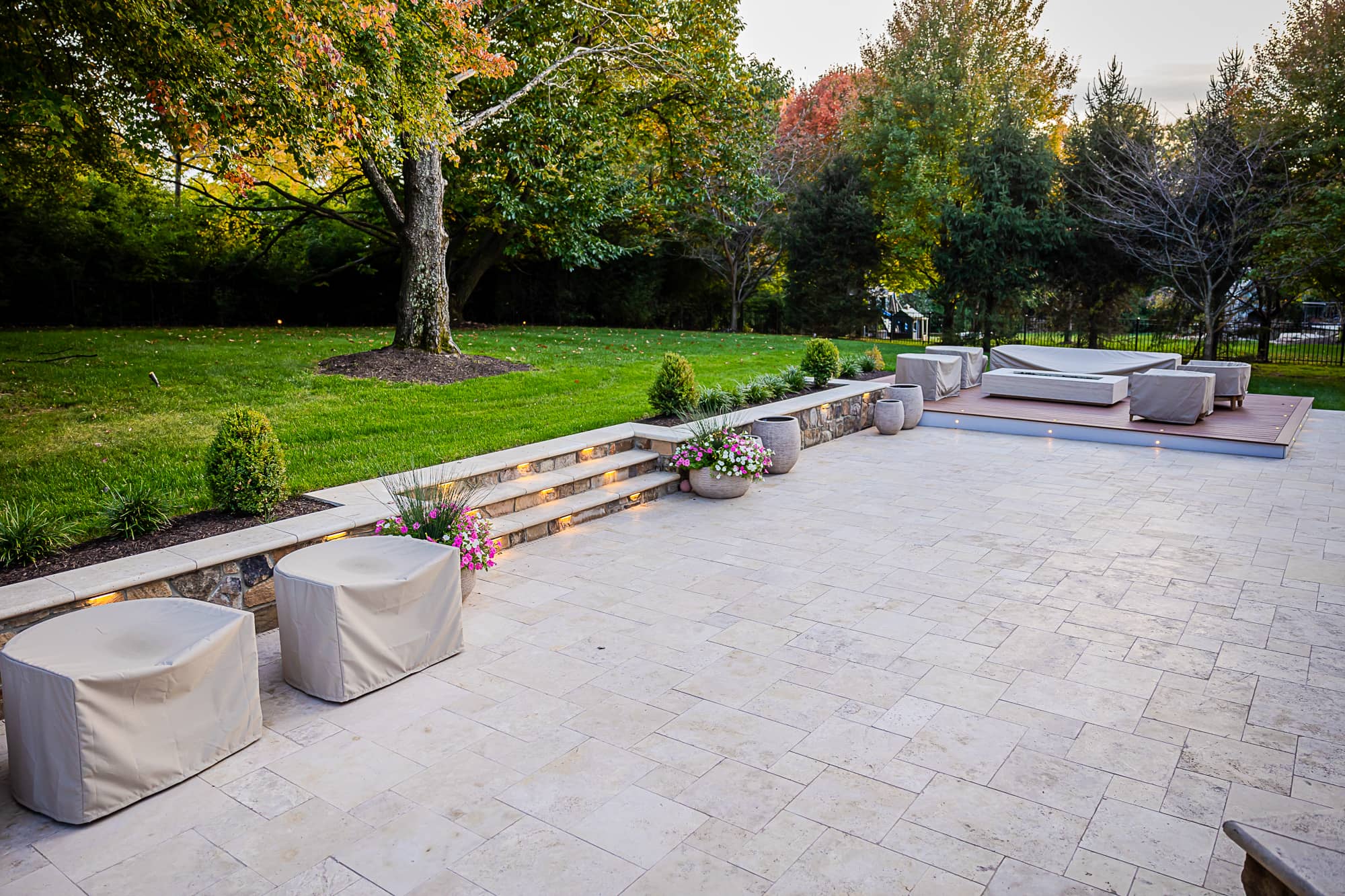 A spacious backyard patio with light stone tiles, part of the Ivory Twilight: A Travertine Patio Project, features covered furniture, potted flowers, and steps with built-in lights, all surrounded by green grass, shrubs, and mature trees in early autumn.