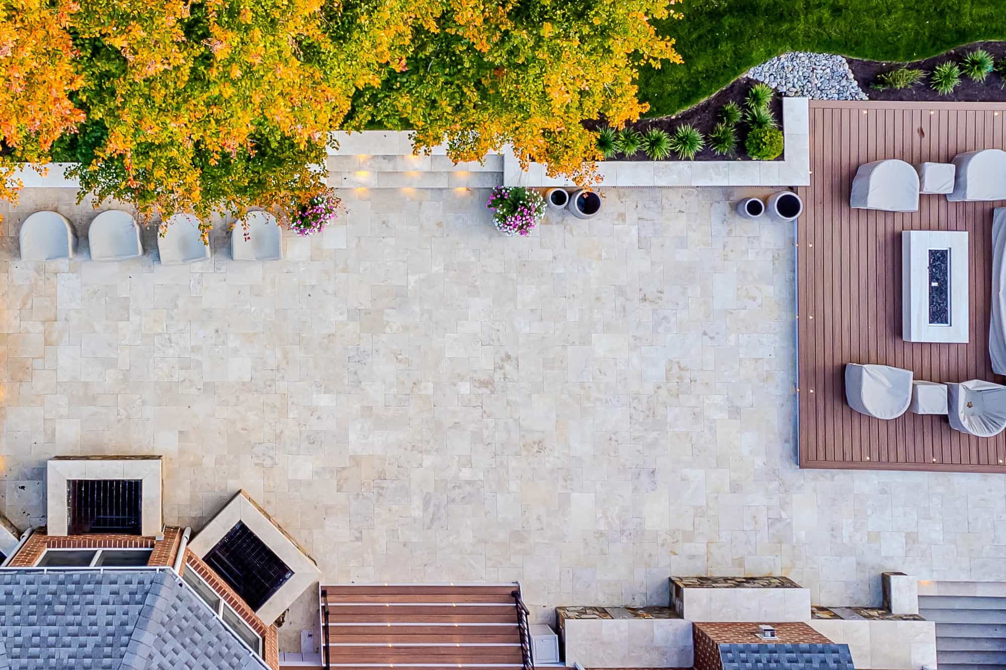 Aerial view of Ivory Twilight: A Travertine Patio Project featuring a stone patio with covered outdoor furniture, fire pit, potted flowers, and lush greenery. A large tree with orange-green leaves casts dappled shade across the area.