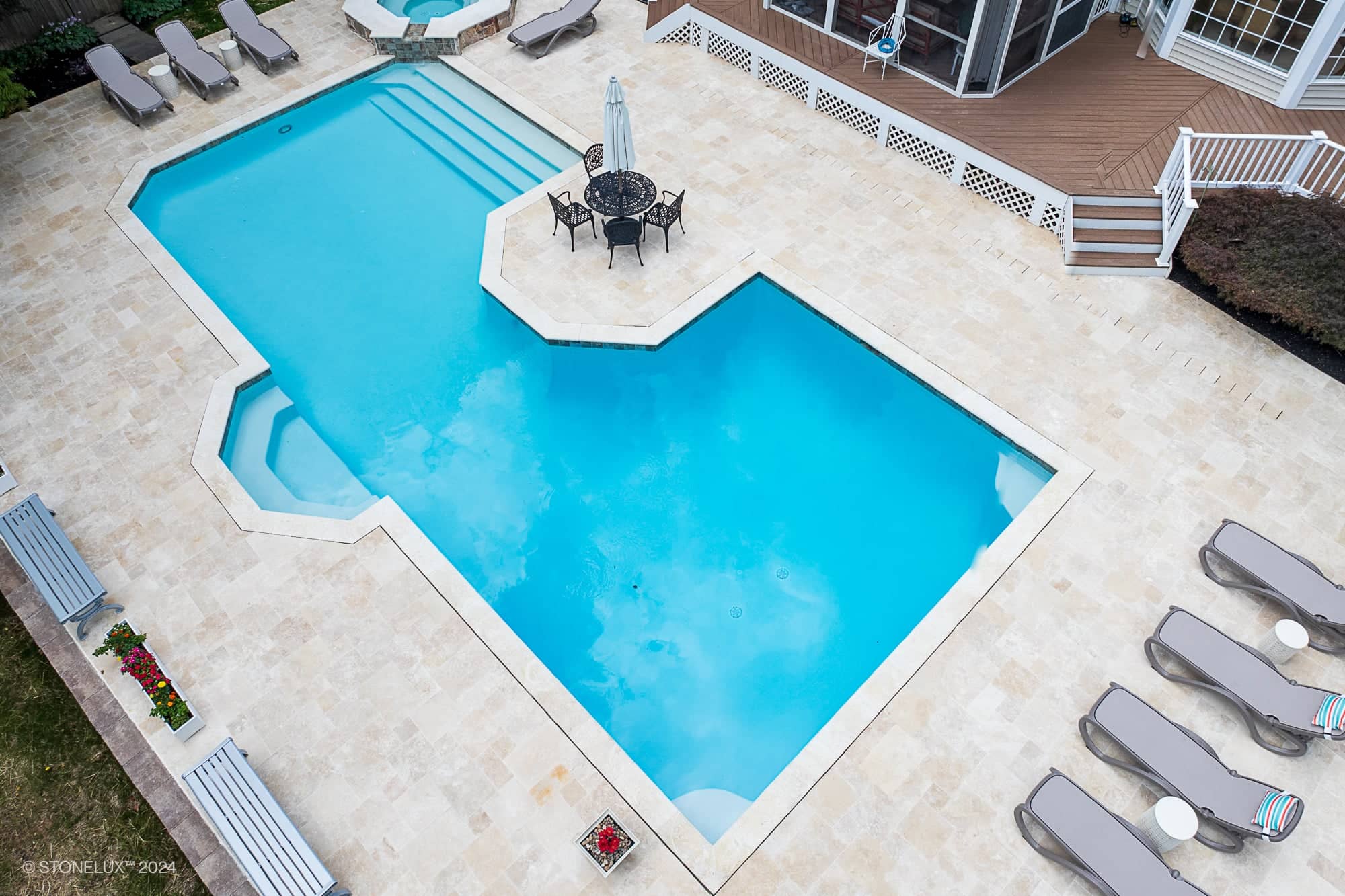 Aerial view of a backyard retreat pool under the trees, featuring a large geometric swimming pool, attached hot tub, ivory travertine tiles, poolside lounge chairs, a round table with chairs, and a deck area near the house.