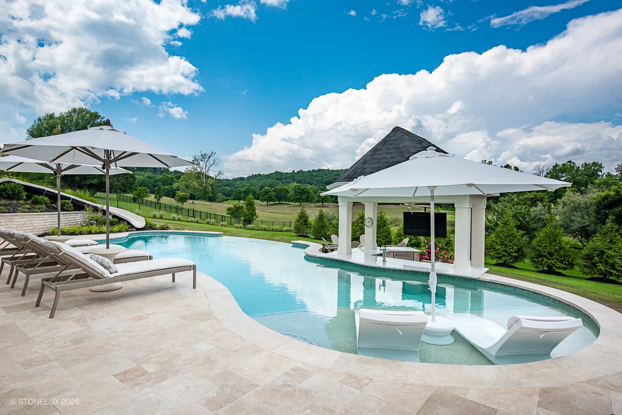 Experience the Leesburg Grecian Oasis – an ivory travertine pool project featuring lounge chairs, umbrellas, a waterslide, and a covered outdoor TV area, all nestled in lush greenery with open field views under a partly cloudy sky.