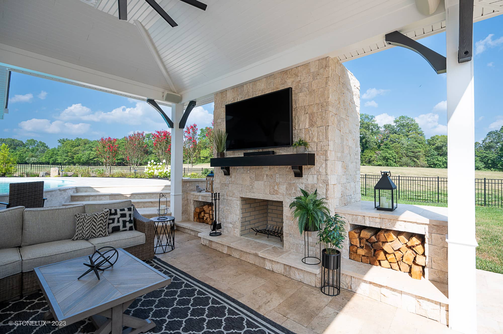 A covered outdoor patio features a stone fireplace with a mounted TV, stacked firewood, and cozy seating, all overlooking a Beige Bliss: Luxurious Travertine Pool and pool house amid trees under a partly cloudy sky.