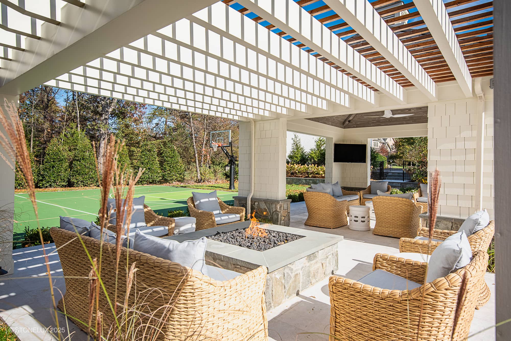 Experience Ashburn Family Fun in this elegant outdoor living space, featuring wicker chairs on Pera Cream Marble under a white pergola. Enjoy the stone firepit, TV, and basketball court—all surrounded by tall trees.