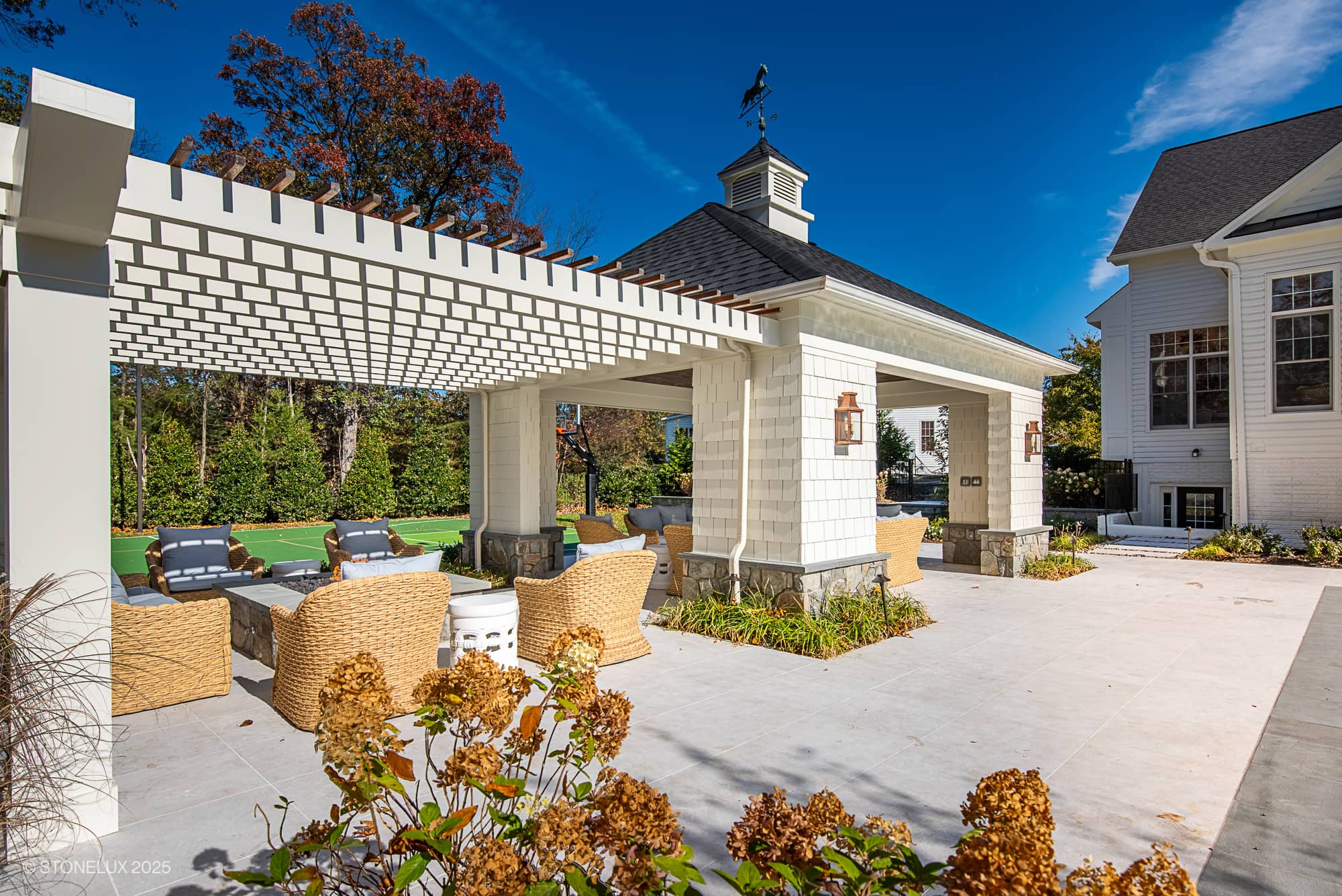 A stylish outdoor lounge area with wicker chairs and sofas under a white pergola, featuring elegant outdoor living in Pera Cream Marble, next to a house and surrounded by greenery, blue sky, and blooming plants in the foreground.