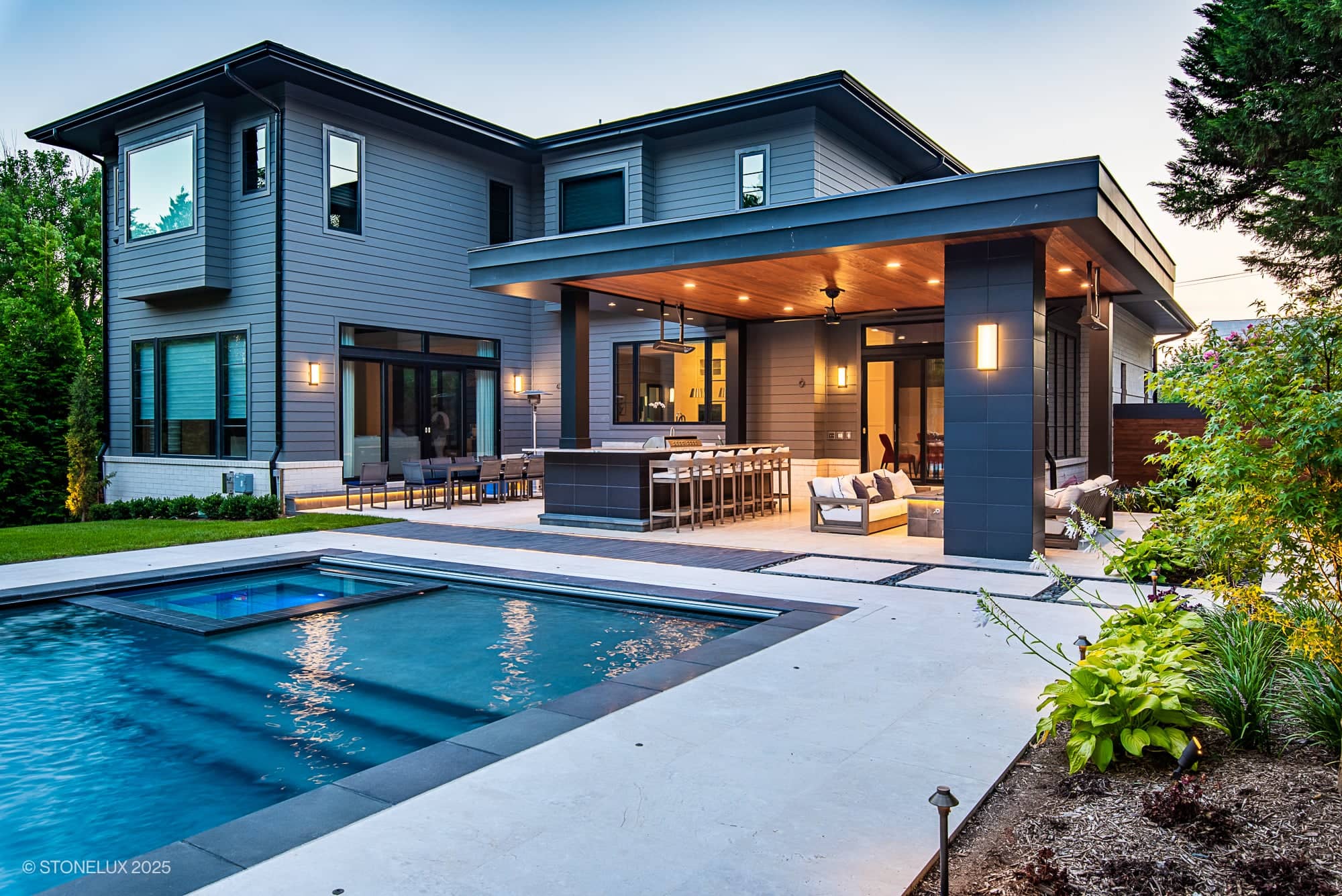 Modern two-story house with gray siding, large windows, and a covered patio featuring an outdoor kitchen and seating area. The McLean Modern Zen design showcases Pera Cream Marble & Black Basalt by the swimming pool and landscaped garden at dusk.