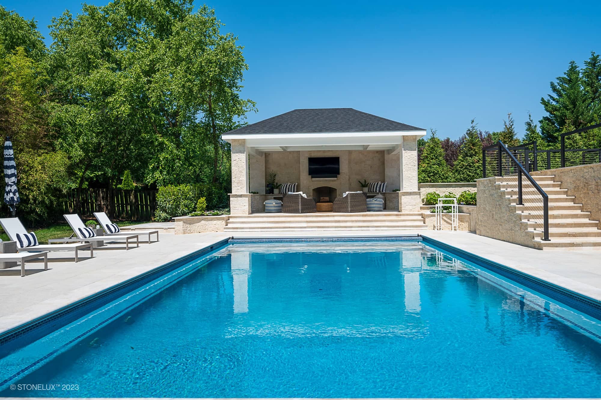 A luxurious backyard pool with clear blue water is surrounded by White Harmony: Pera Cream Sandblasted Marble lounge chairs and umbrellas, facing a porcelain poolside retreat with a TV and greenery beneath a bright blue sky.
