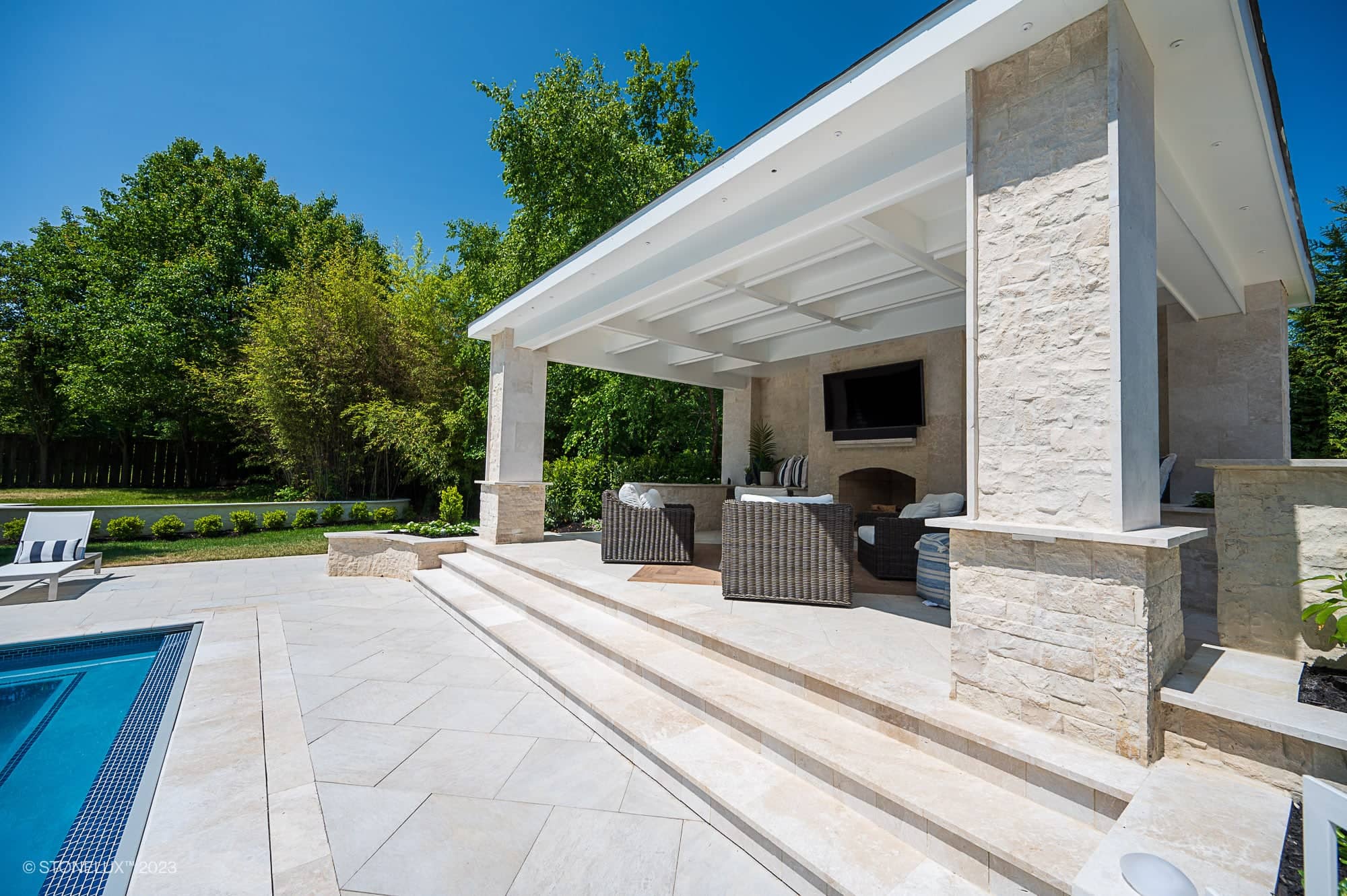 A modern outdoor patio with White Harmony: Pera Cream Sandblasted Marble steps leading to a covered seating area, wicker furniture, and a mounted TV. The porcelain poolside retreat overlooks a pool and lush greenery under a clear blue sky.