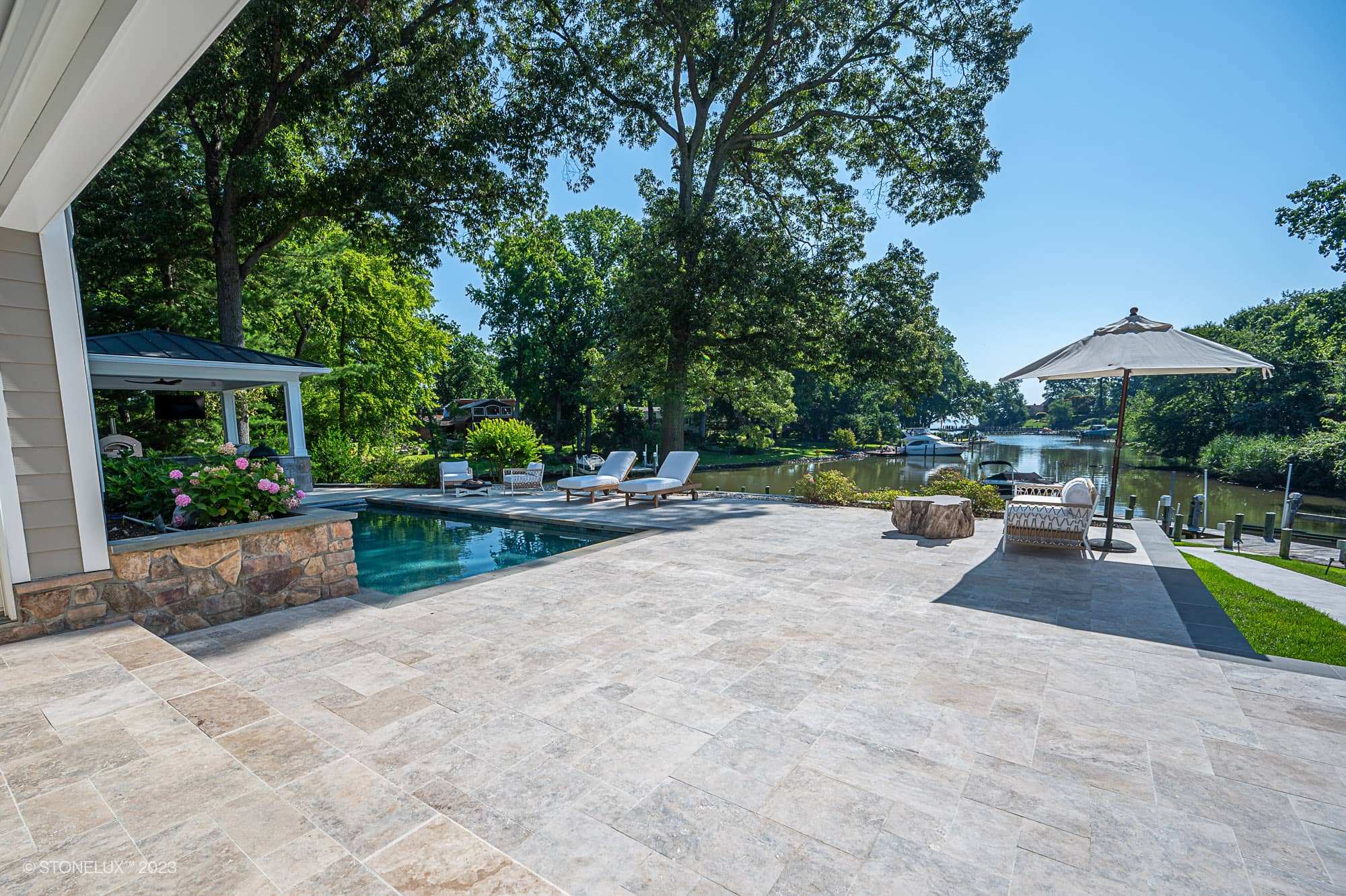 A spacious stone patio with lounge chairs, an umbrella, and a swimming pool—part of the Annapolis Dream – Philly Travertine Pool Project—overlooks a scenic river surrounded by lush green trees and a clear blue sky.