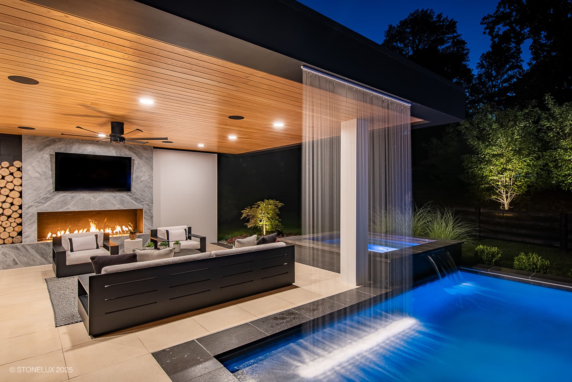 Aldie Modern Escape – Porcelain & Basalt Luxury Pool Pavilion: modern outdoor living area at night with a pool, waterfall feature, lounge seating, fireplace, mounted TV, and stacked firewood; illuminated trees and serene ambiance complete the scene.