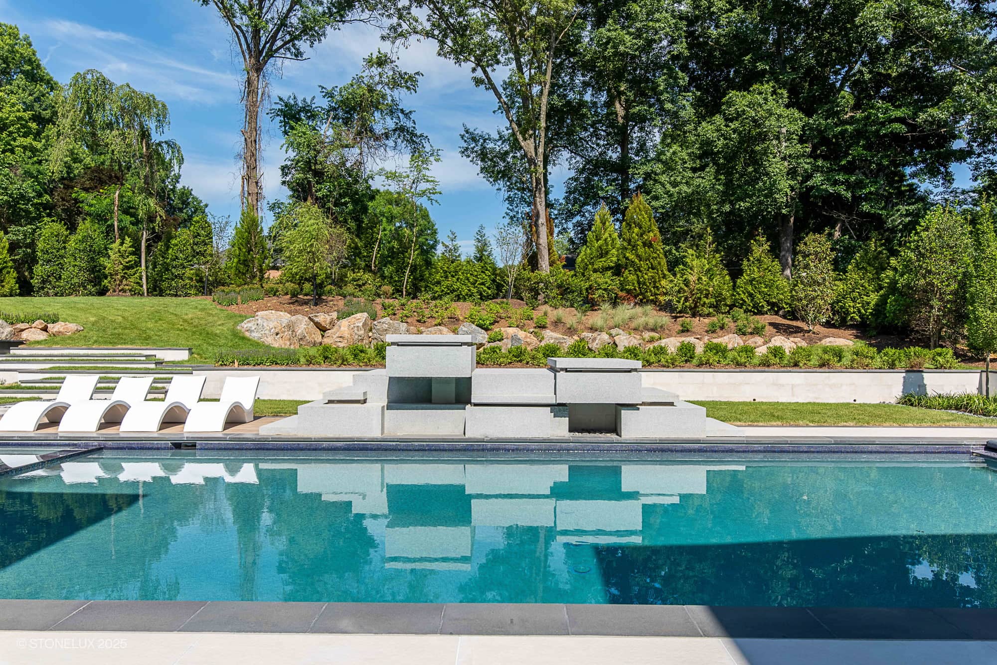 A modern outdoor pool with clear blue water, surrounded by white lounge chairs and sleek stone structures. This Modern Hillside Elegance – McLean Retreat features Overland Pearl Porcelain and Black Basalt amid lush greenery.