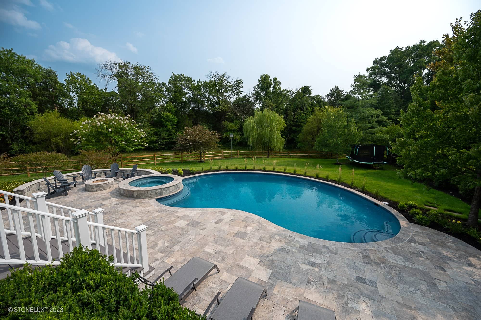 Experience the Silver Splash: The Silver Travertine Pool Haven—a backyard retreat featuring a curved swimming pool and hot tub, stone patio with lounge chairs and firepit, all bordered by lush grass, trees, and a wooden fence.