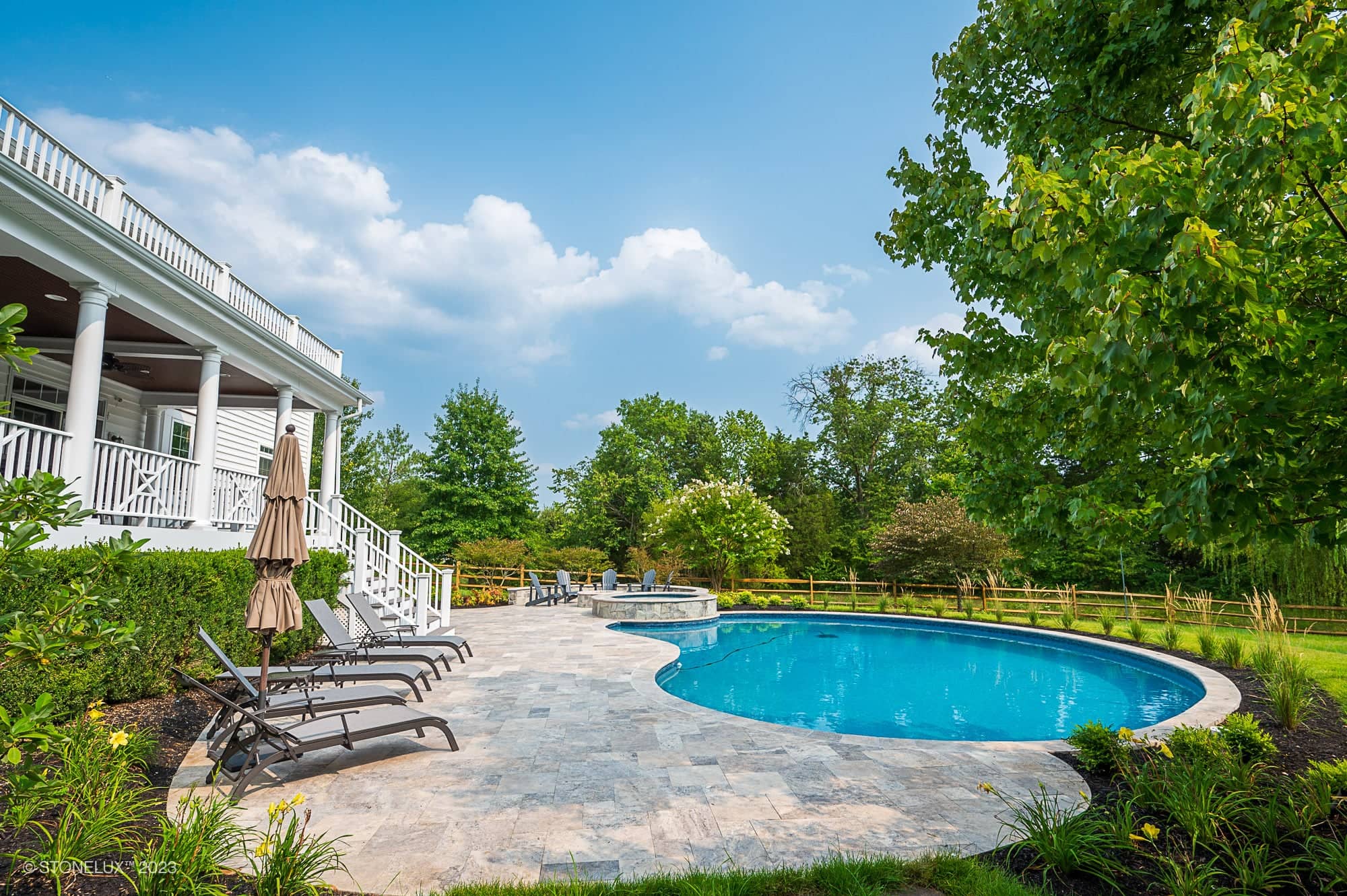 A backyard scene featuring Silver Splash: The Silver Travertine Pool Haven—a curved swimming pool framed by a stone patio, lounge chairs with umbrellas, lush green trees, and a white two-story house with a covered porch under blue skies.