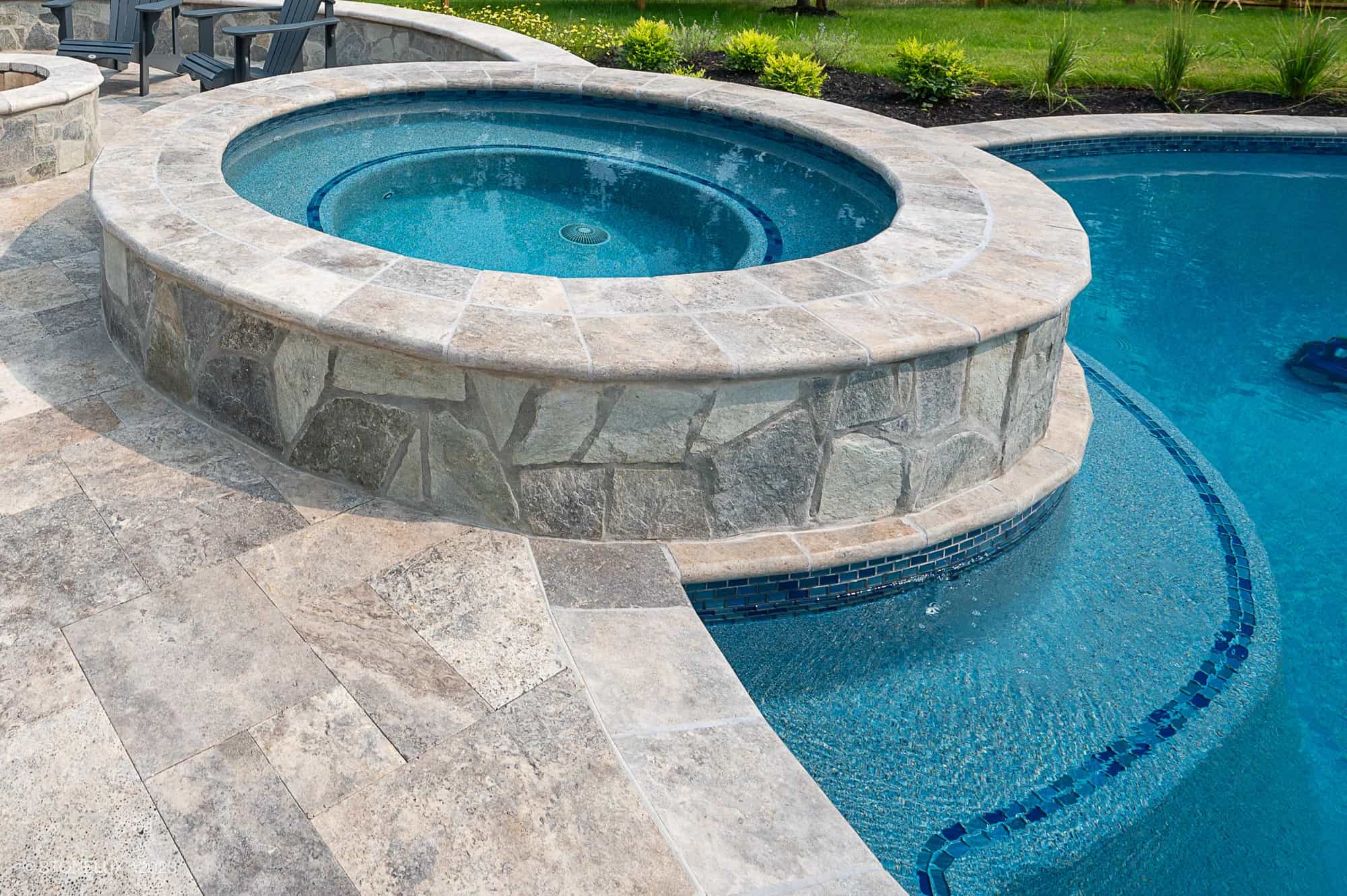 A round raised hot tub with stone tiles is connected to a larger swimming pool, creating the Silver Splash: The Silver Travertine Pool Haven. Light-colored stone decking surrounds the area, accented by green plants in the background.