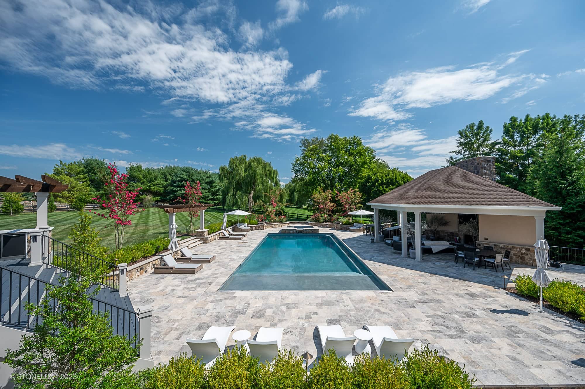 A luxurious outdoor pool area, Silver Expanse: The Silver Travertine Pool Landscape features lounge chairs, umbrellas, and a covered patio surrounded by landscaped greenery and trees under a bright blue sky with scattered clouds.