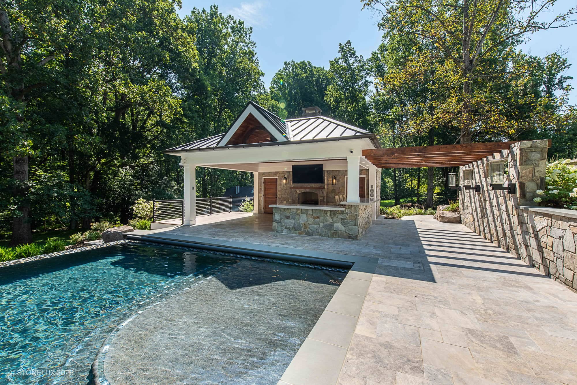 A modern backyard pool at the Clifton Cascade Retreat – Silver Travertine Oasis features a stone patio, pool house, shaded pergola, and lush green trees surrounding the area on a sunny day.