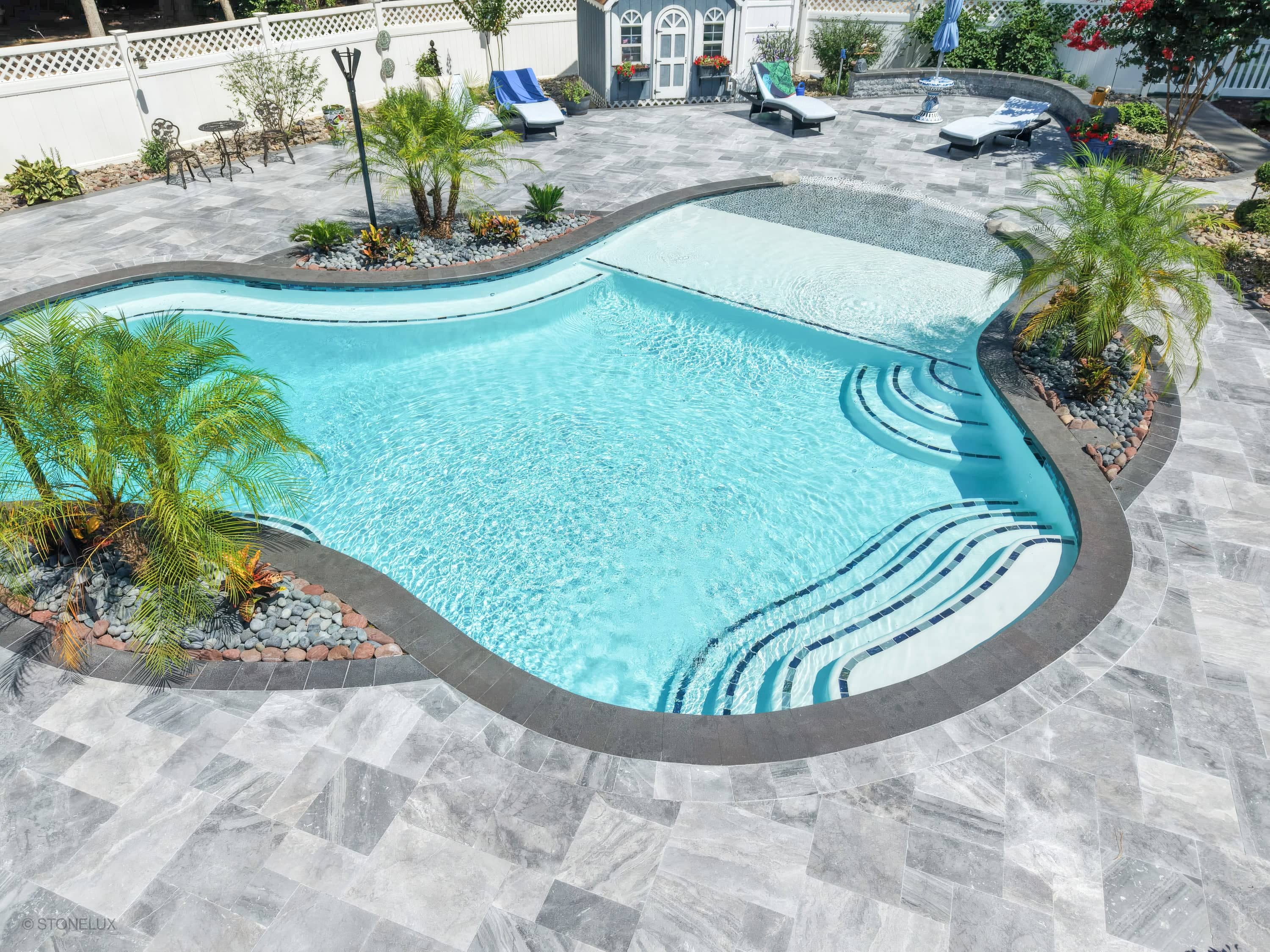 A modern backyard oasis in Salisbury, MD, featuring the Aegean Grey Marble Pool Retreat with sleek black basalt coping. The shimmering blue pool water complements the grey marble pavers and lush tropical landscaping. Elegant lounge chairs and a stylish...
