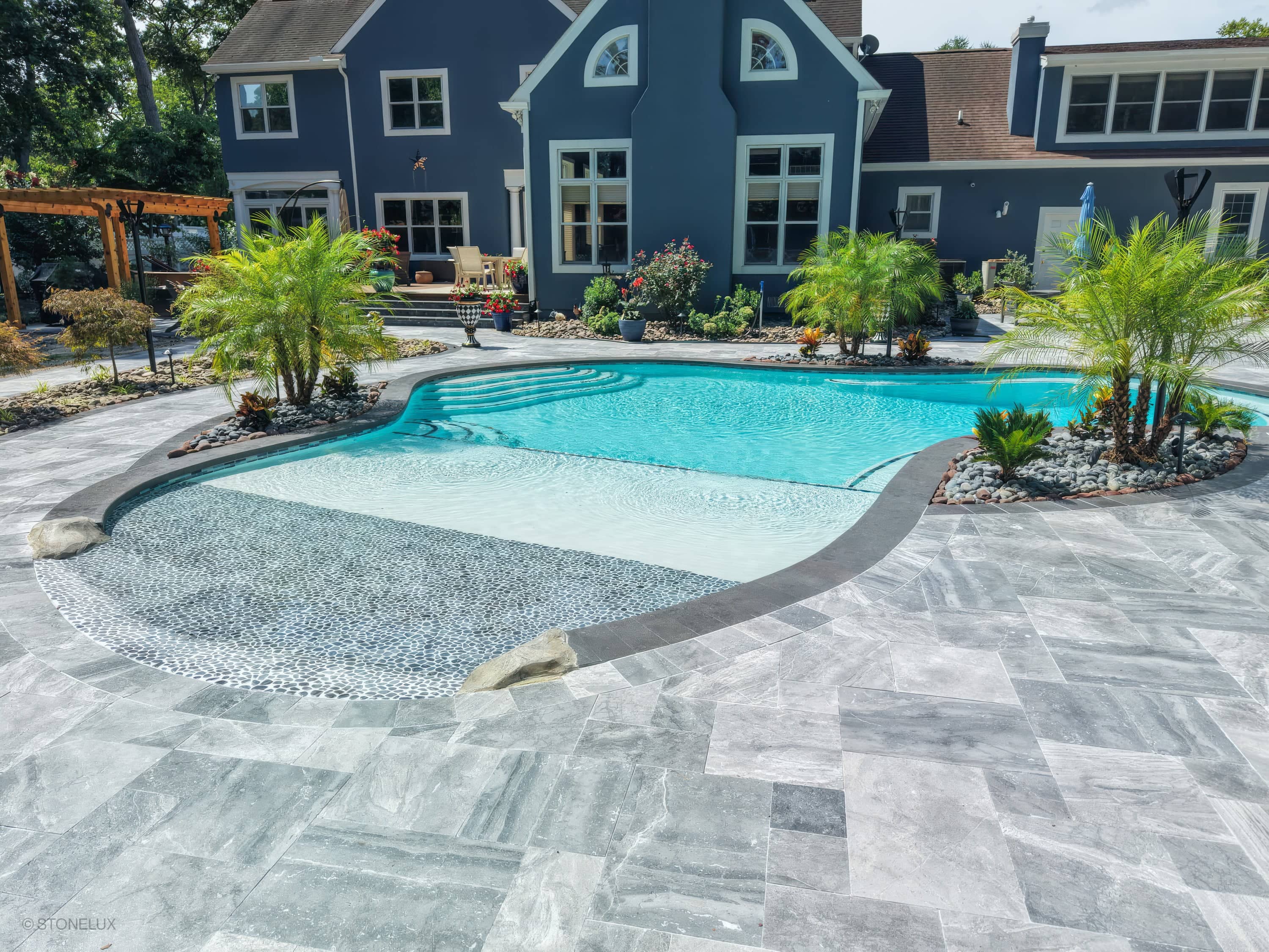 Aegean Grey Marble Pool Retreat in Salisbury, MD features a sleek modern design with black basalt coping, shimmering blue water, lush palm landscaping, and elegant grey pavers. The luxurious pool area exudes a serene ambiance, perfect for a sophisticat...