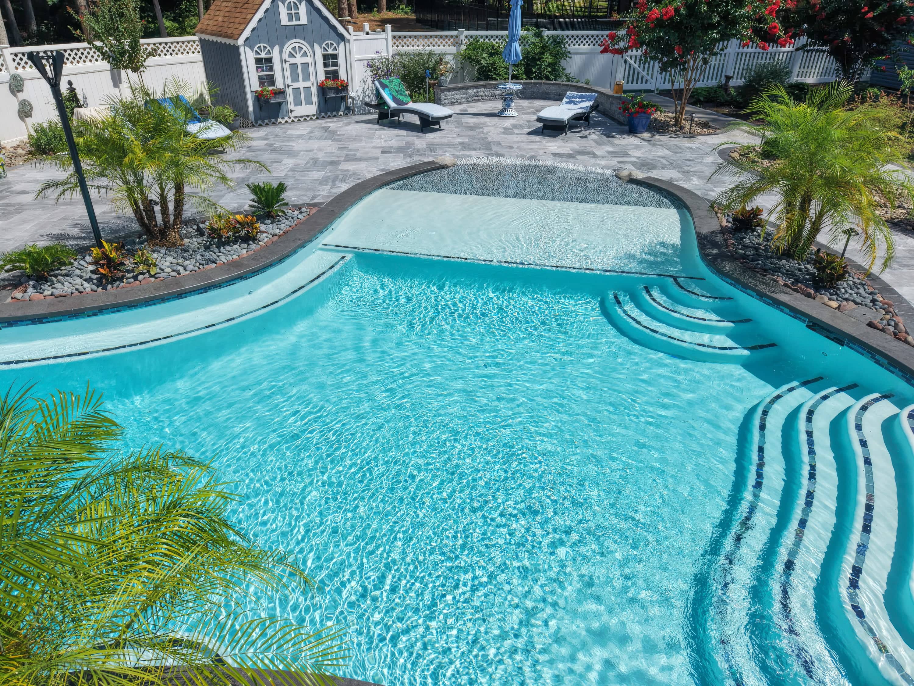 Aegean Grey Marble Pool Retreat in Salisbury, MD, features sleek black basalt coping. The modern design includes clear blue water, curved steps, and lush tropical landscaping. The grey patio harmonizes with white loungers and vibrant plants, creating a...