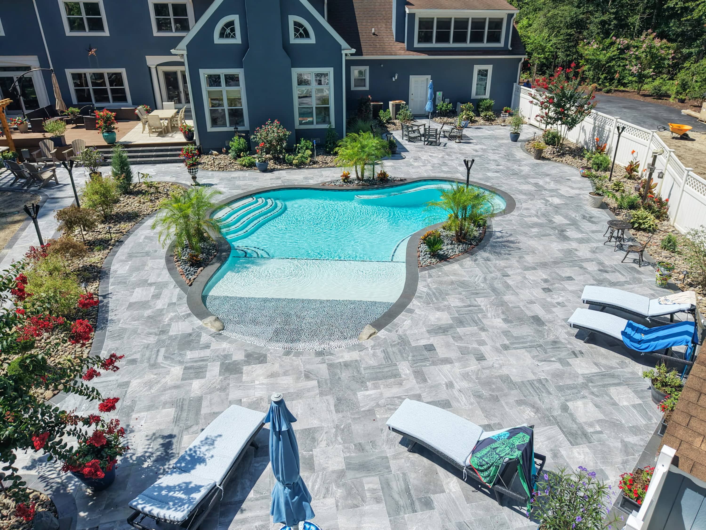 Aegean Grey Marble Pool Retreat in Salisbury, MD, features sleek grey marble pavers and black basalt coping. The modern design surrounds a sparkling blue pool, complemented by lush landscaping, red blooms, and white lounge chairs, creating a serene bac...