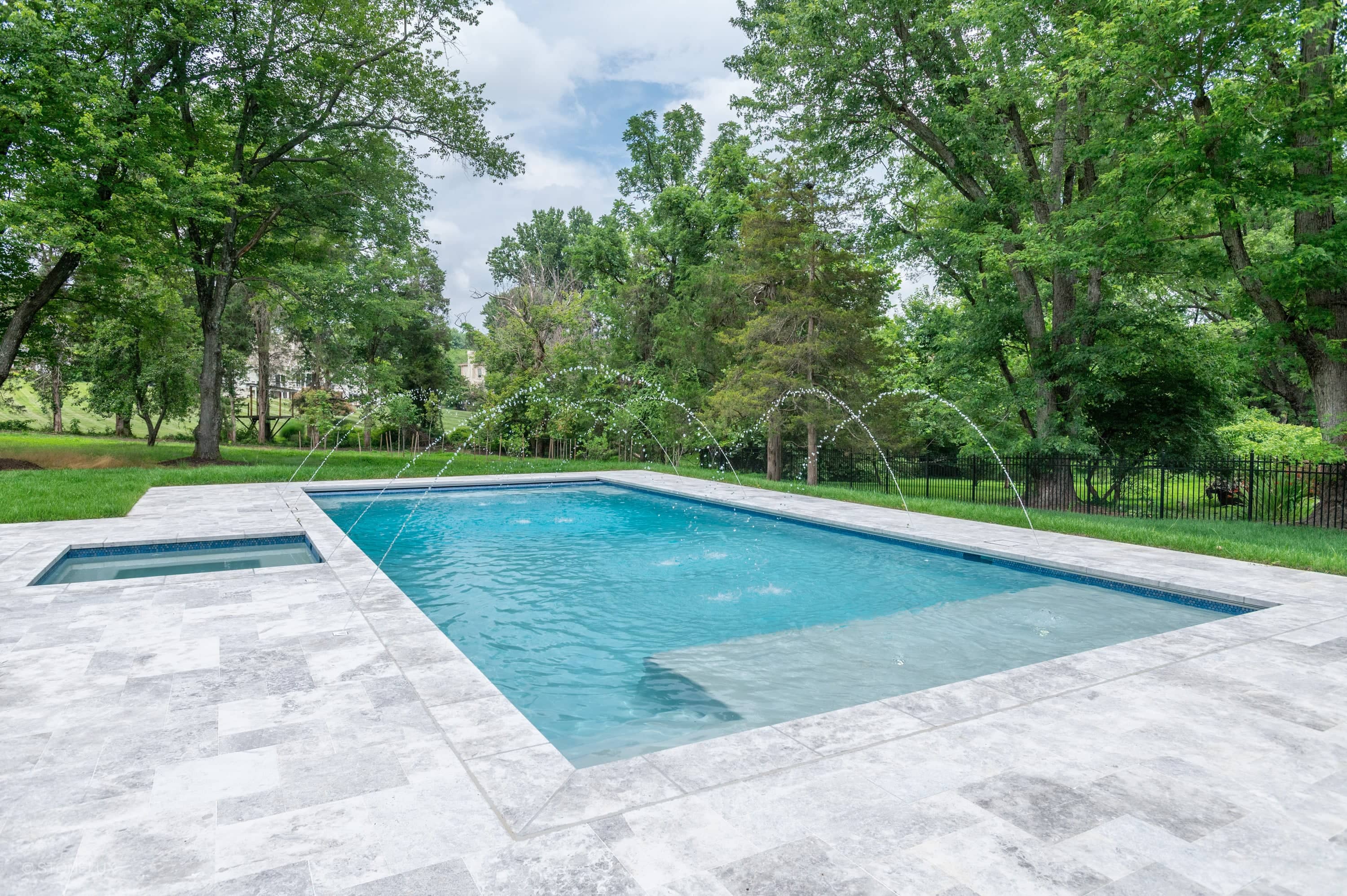 Tundra Grey Harmony marble pool retreat in McLean, VA, features elegant marble coping and pavers. The serene blue pool is surrounded by lush greenery, creating a modern oasis. Water features enhance tranquility, while the natural stone complements the ...