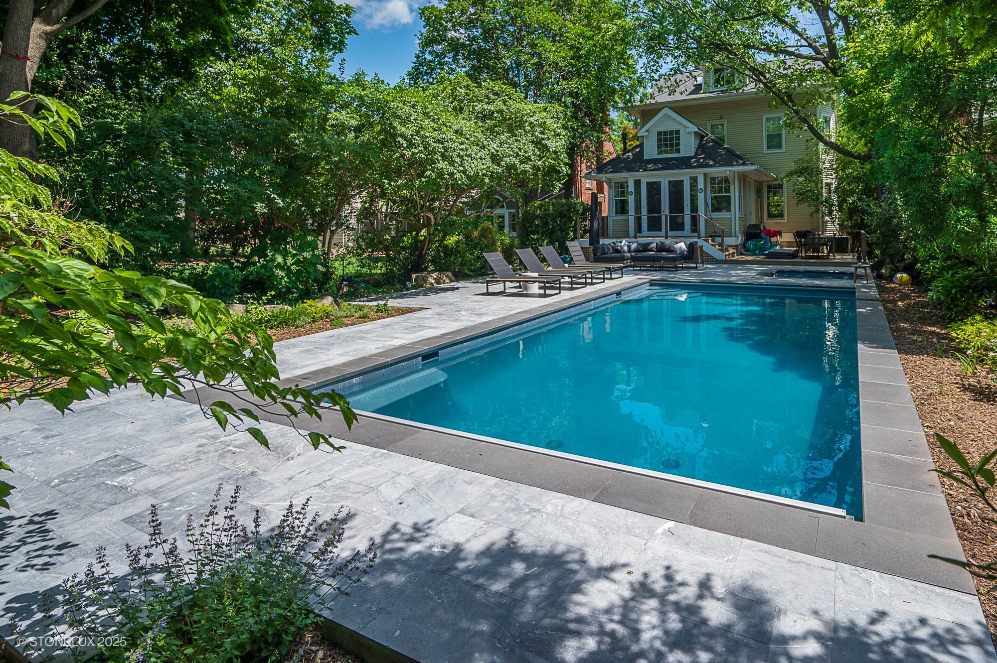 Experience the Urban Serenity Pool Project—Aegean Grey Marble & Black Basalt accent this rectangular backyard pool with clear blue water, surrounded by a stone deck, lounge chairs, lush trees, and a two-story house with a screened porch.