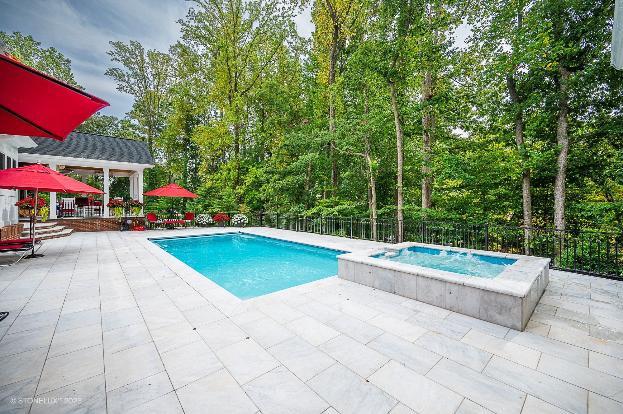 A spacious backyard features a rectangular swimming pool crafted with White Whisper: The Pristine Afyon Marble Pool Elegance, an adjacent hot tub, and a white stone patio with red umbrellas, lounge chairs, and lush green trees in the background.