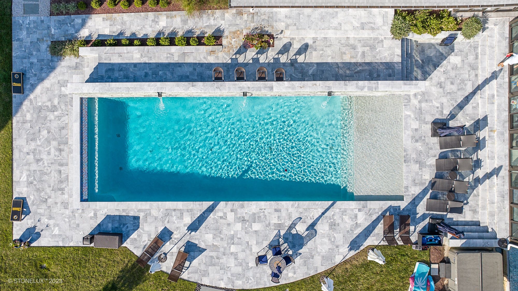 A rectangular outdoor swimming pool, part of the Blue Haven: The Grey and Blue Sandblasted Marble Pool Project, is surrounded by a stone deck, lounge chairs, and greenery. Three people are in the pool; umbrellas and seating are arranged around the deck.