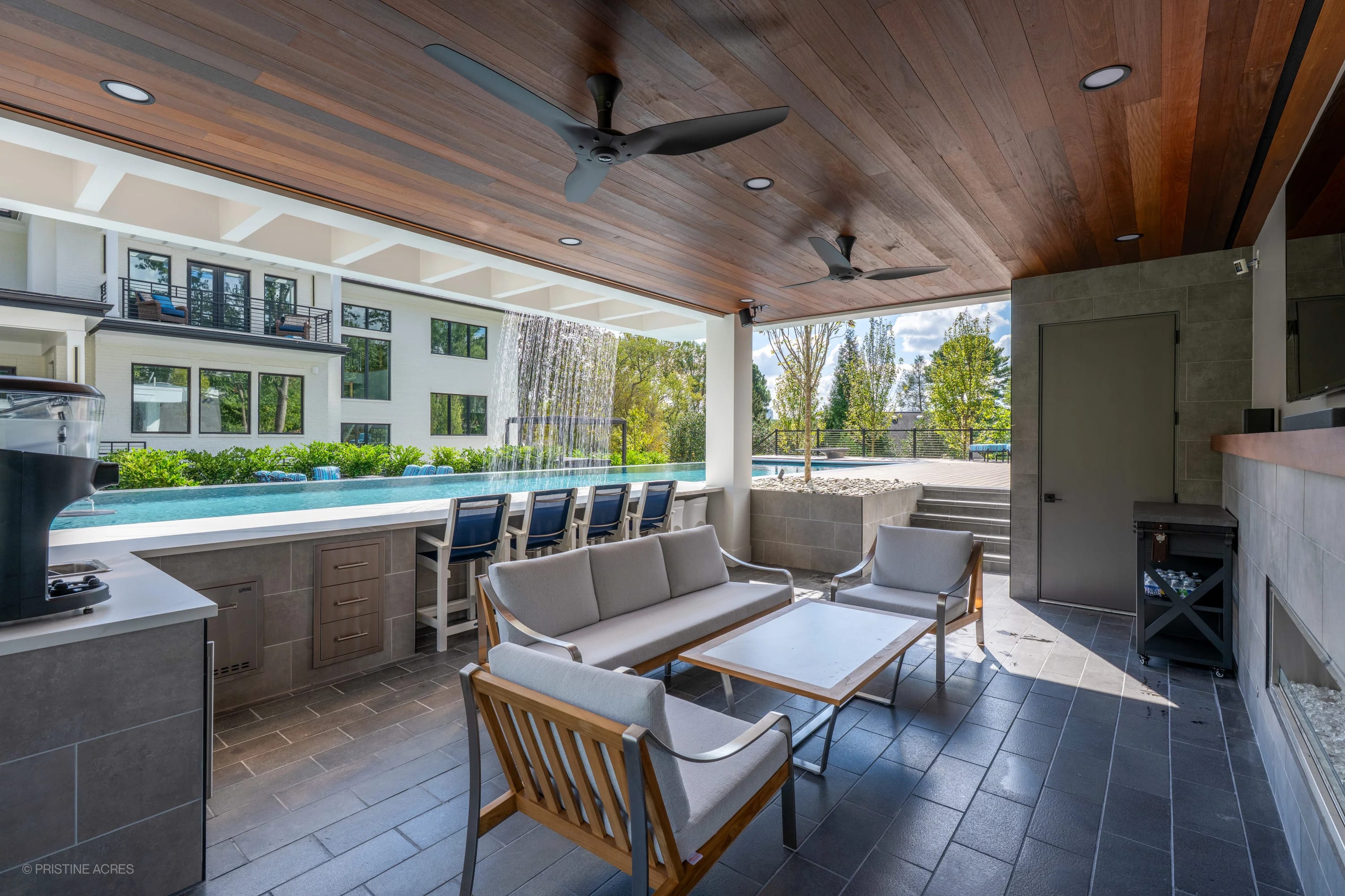 Modern McLean Pool Project in McLean, VA features Black Basalt flooring and Nexus White Porcelain. The sleek patio includes a cascading water feature, grey outdoor furniture, and lush greenery, creating a contemporary, serene backyard oasis.