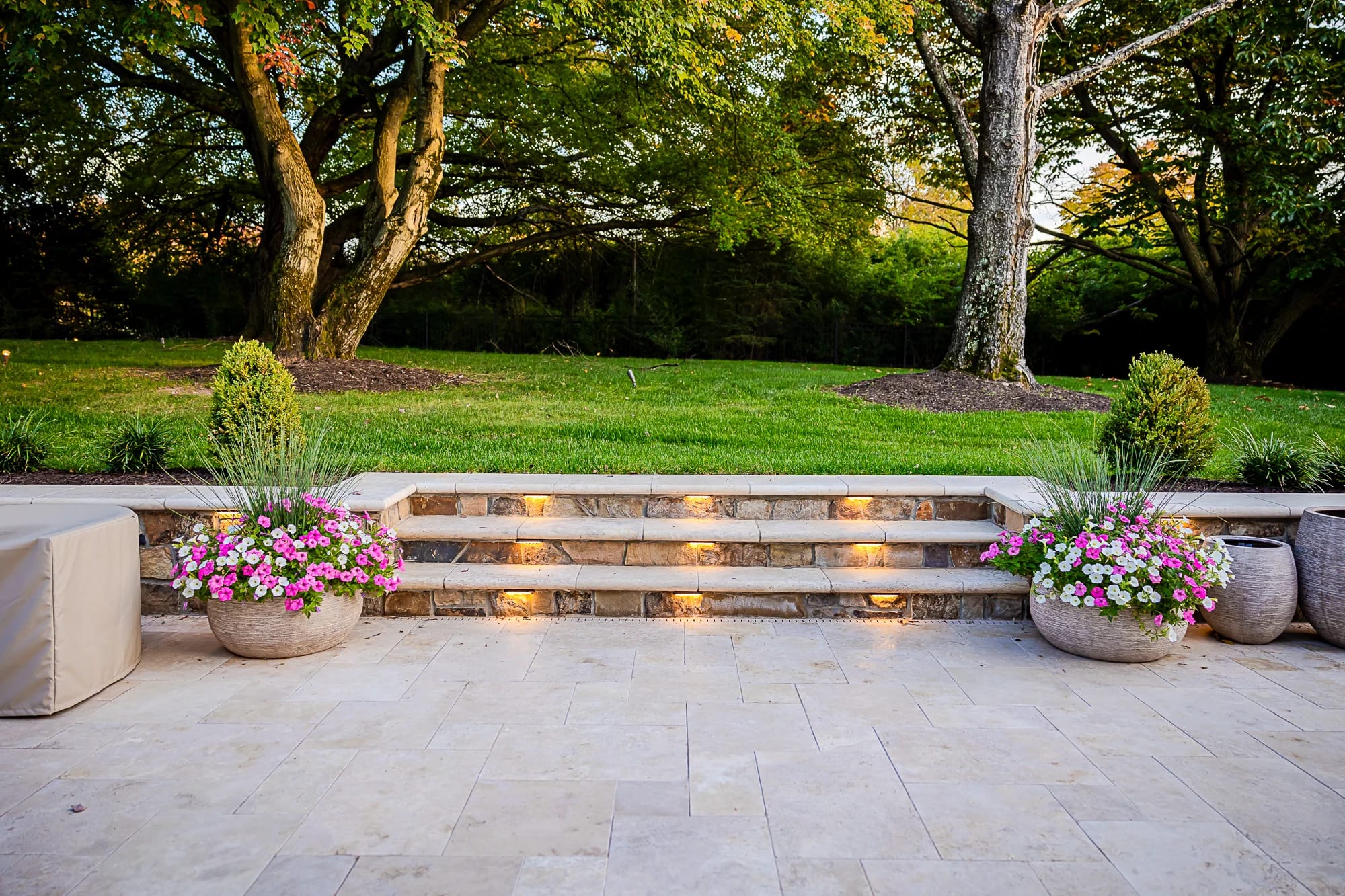 Ivory Twilight: A Travertine Patio Project features stone steps with built-in lights leading to a green lawn bordered by trees. Large planters with colorful flowers and tall grass flank the steps, creating a serene outdoor setting.