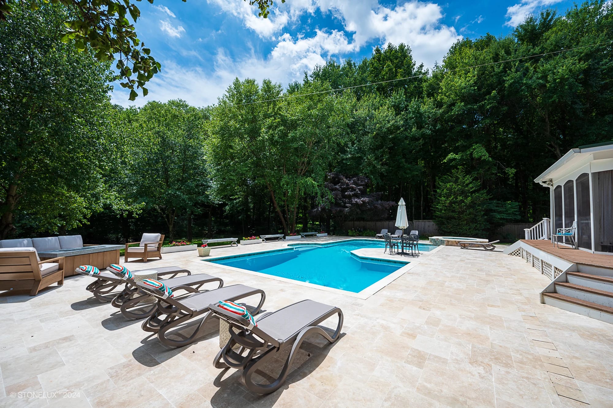A sunny backyard features a rectangular swimming pool with Ivory Travertine decking—your retreat pool under the trees—surrounded by lounge chairs, umbrellas, and seating areas, with lush greenery and a clear blue sky overhead.