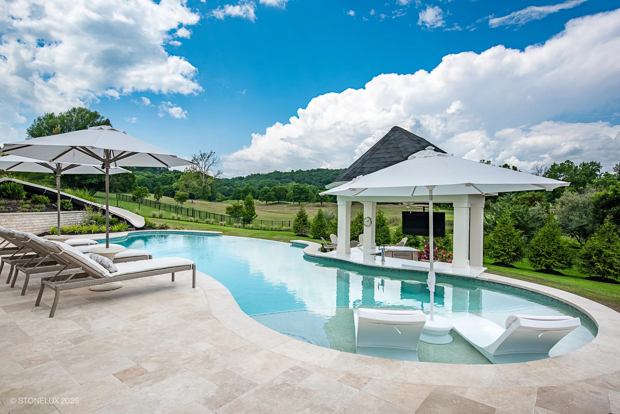 Experience the Leesburg Grecian Oasis – an ivory travertine pool project featuring lounge chairs, umbrellas, a waterslide, and a covered outdoor TV area, all nestled in lush greenery with open field views under a partly cloudy sky.