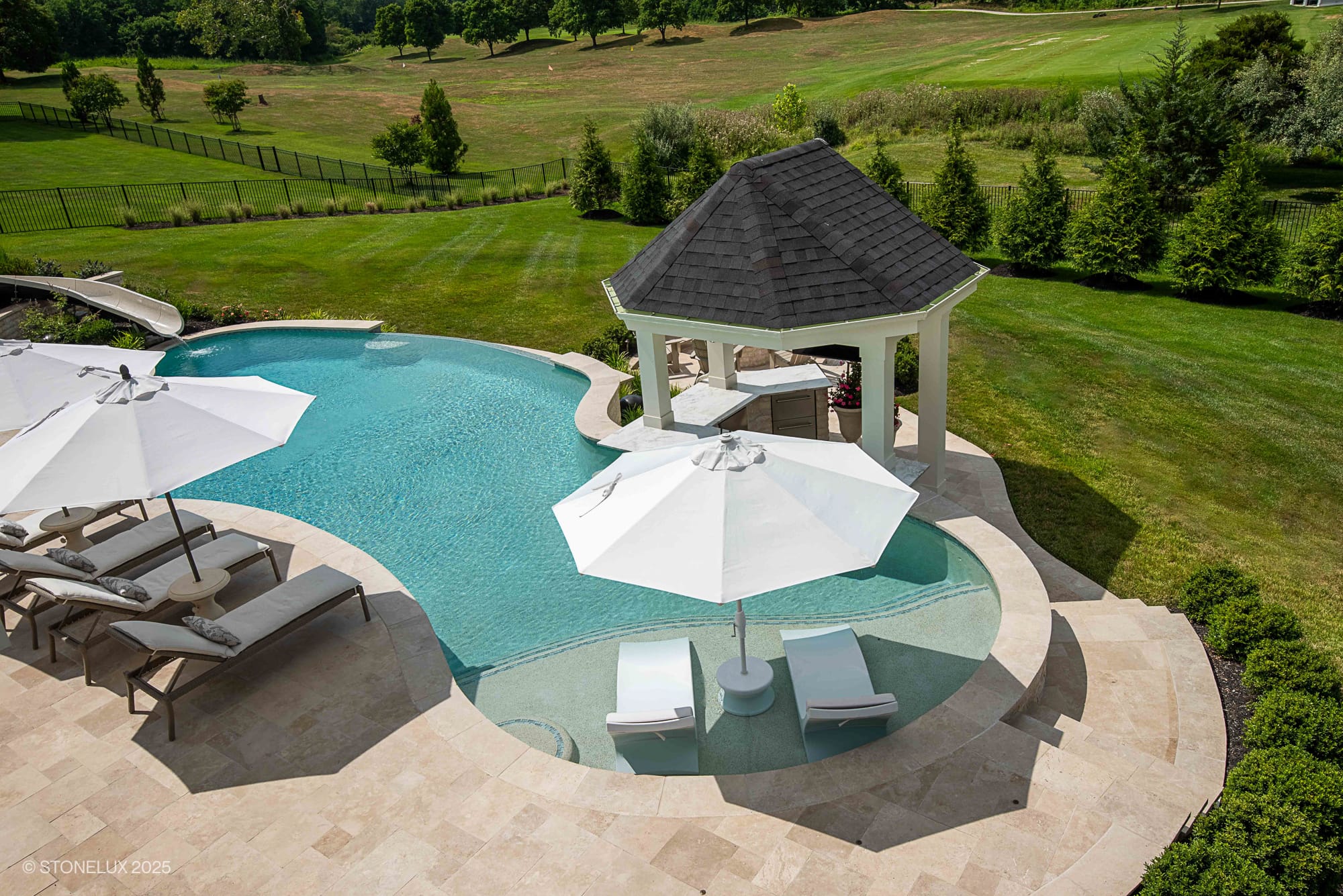 A luxurious Leesburg Grecian Oasis featuring an ivory travertine pool with sun loungers under white umbrellas, a shaded pavilion, and a waterslide, all surrounded by green lawn and trees on a sunny day.