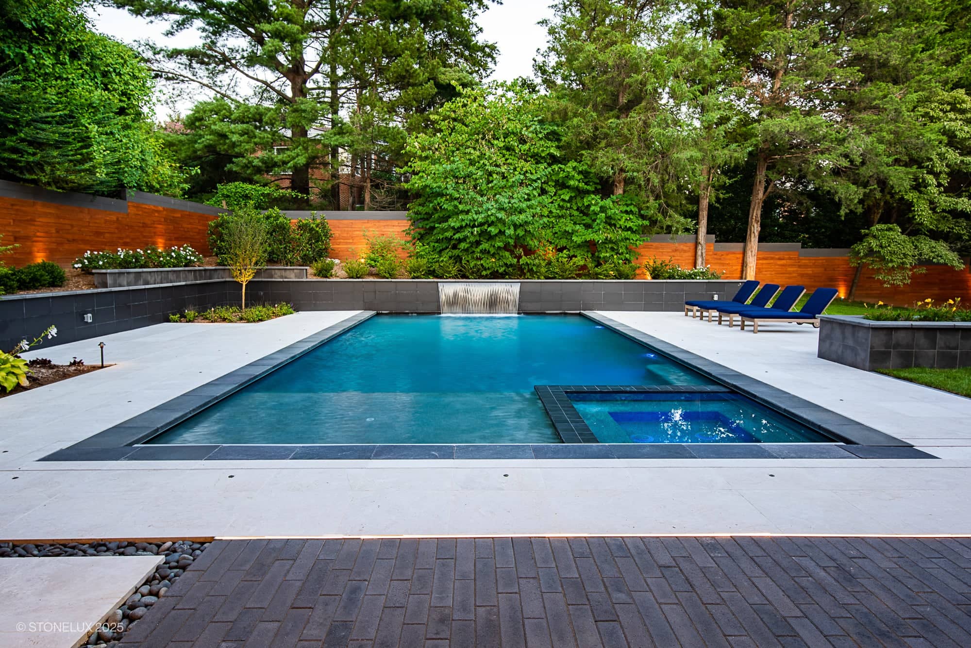 A modern backyard with a rectangular swimming pool, attached hot tub, and waterfall feature creates a serene escape. Blue lounge chairs line the patio, while McLean Modern Zen – Outdoor Living in Pera Cream Marble & Black Basalt enhances the scene.