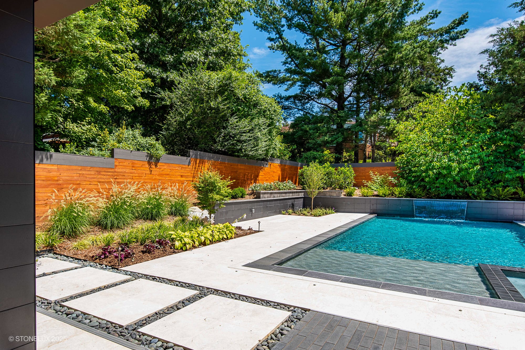 A modern backyard with a swimming pool, lush green trees, and wooden fencing. The patio showcases McLean Modern Zen – Outdoor Living in Pera Cream Marble & Black Basalt, complemented by a small waterfall and neat landscaping.