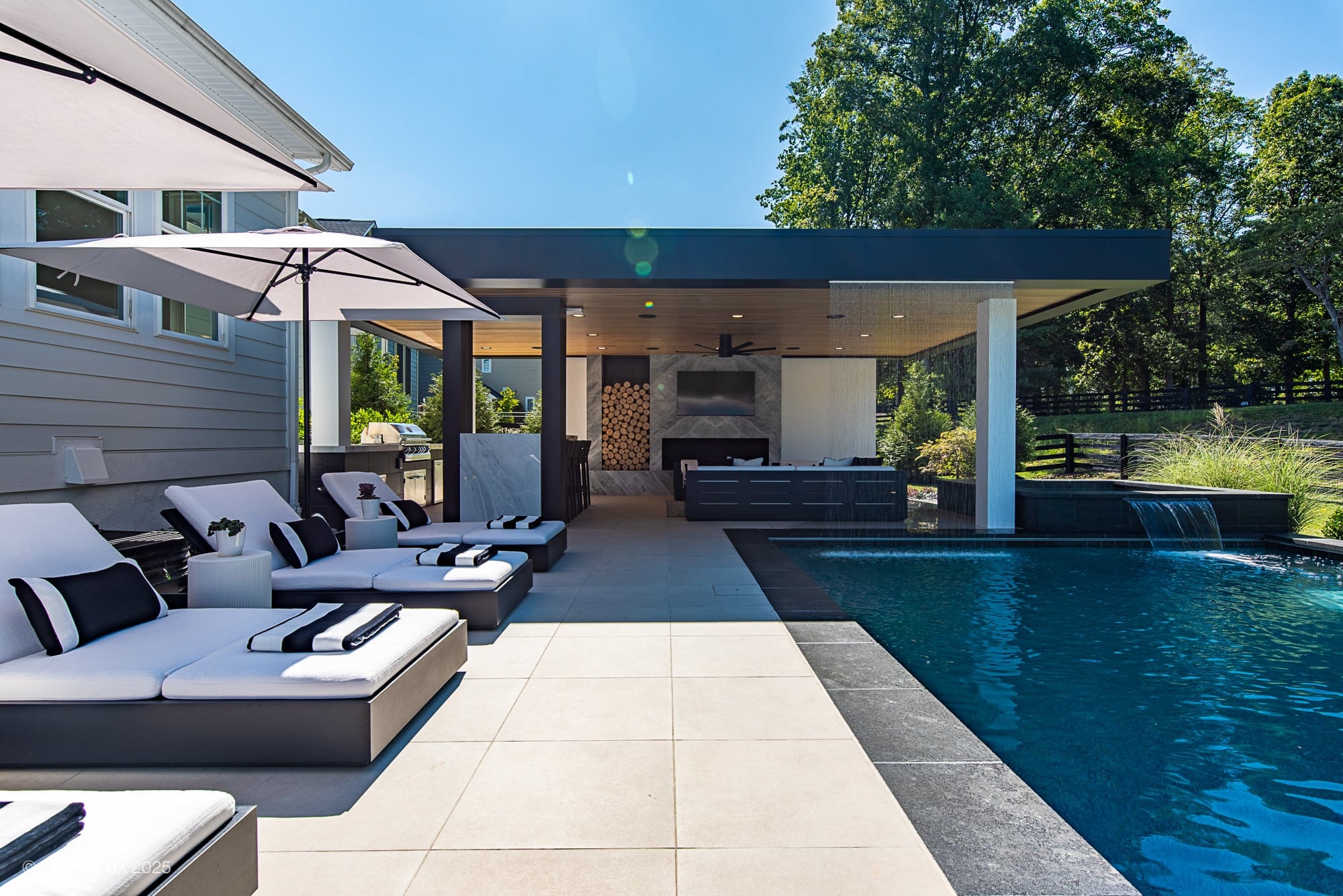Aldie Modern Escape – Porcelain & Basalt Luxury Pool Pavilion: Modern backyard with a pool, lounge chairs with umbrellas, and a covered patio area featuring a fireplace, outdoor kitchen. Lush trees and blue sky complete the serene retreat.