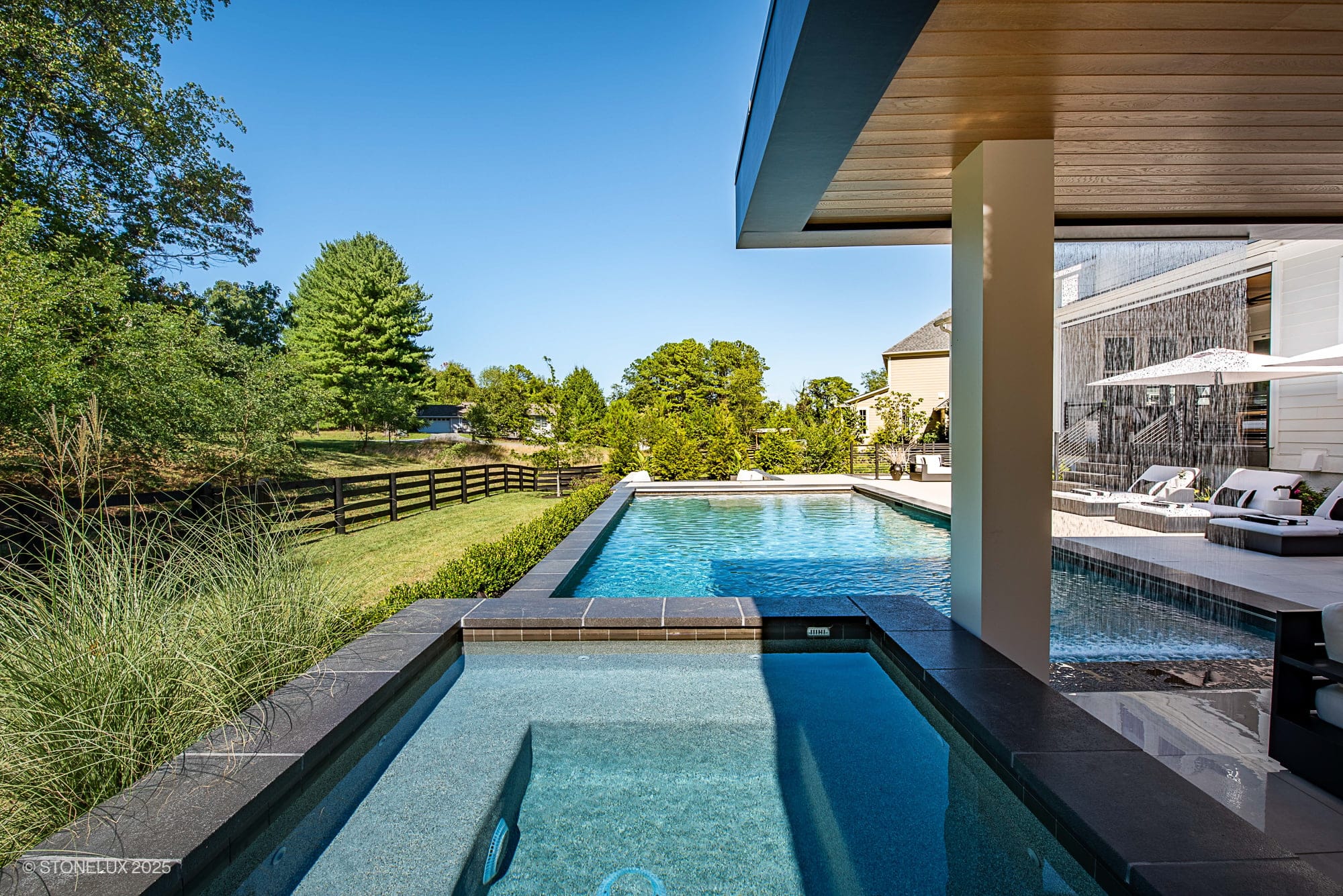 Aldie Modern Escape – Porcelain & Basalt Luxury Pool Pavilion: This modern backyard features a rectangular pool and hot tub, surrounded by greenery and a wooden fence, with lounge chairs and a covered patio on a clear, sunny day.