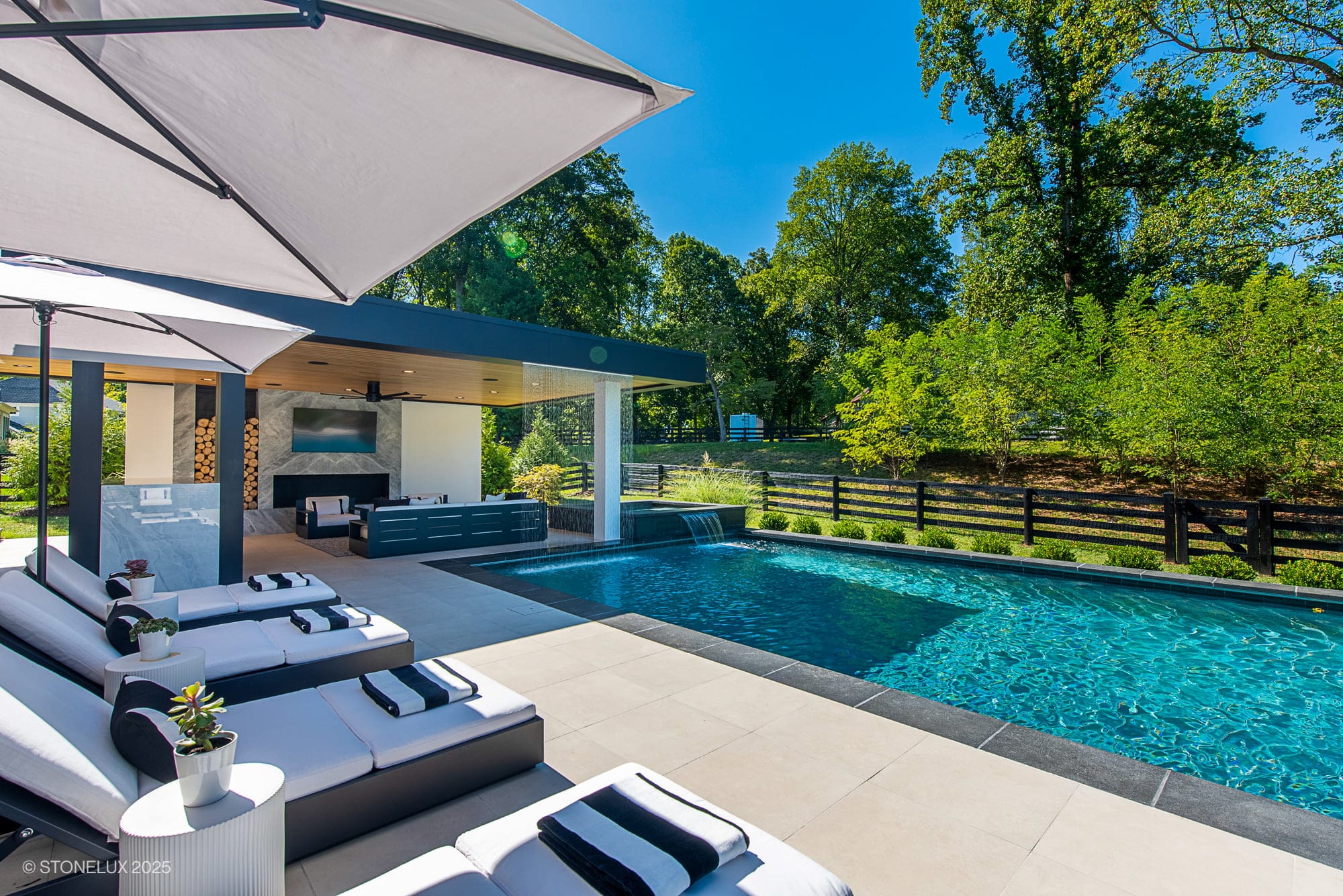Experience the Aldie Modern Escape – a Porcelain & Basalt Luxury Pool Pavilion featuring lounge chairs, an umbrella, a covered seating space, waterfall feature, and lush green trees framing this modern outdoor oasis on a sunny day.