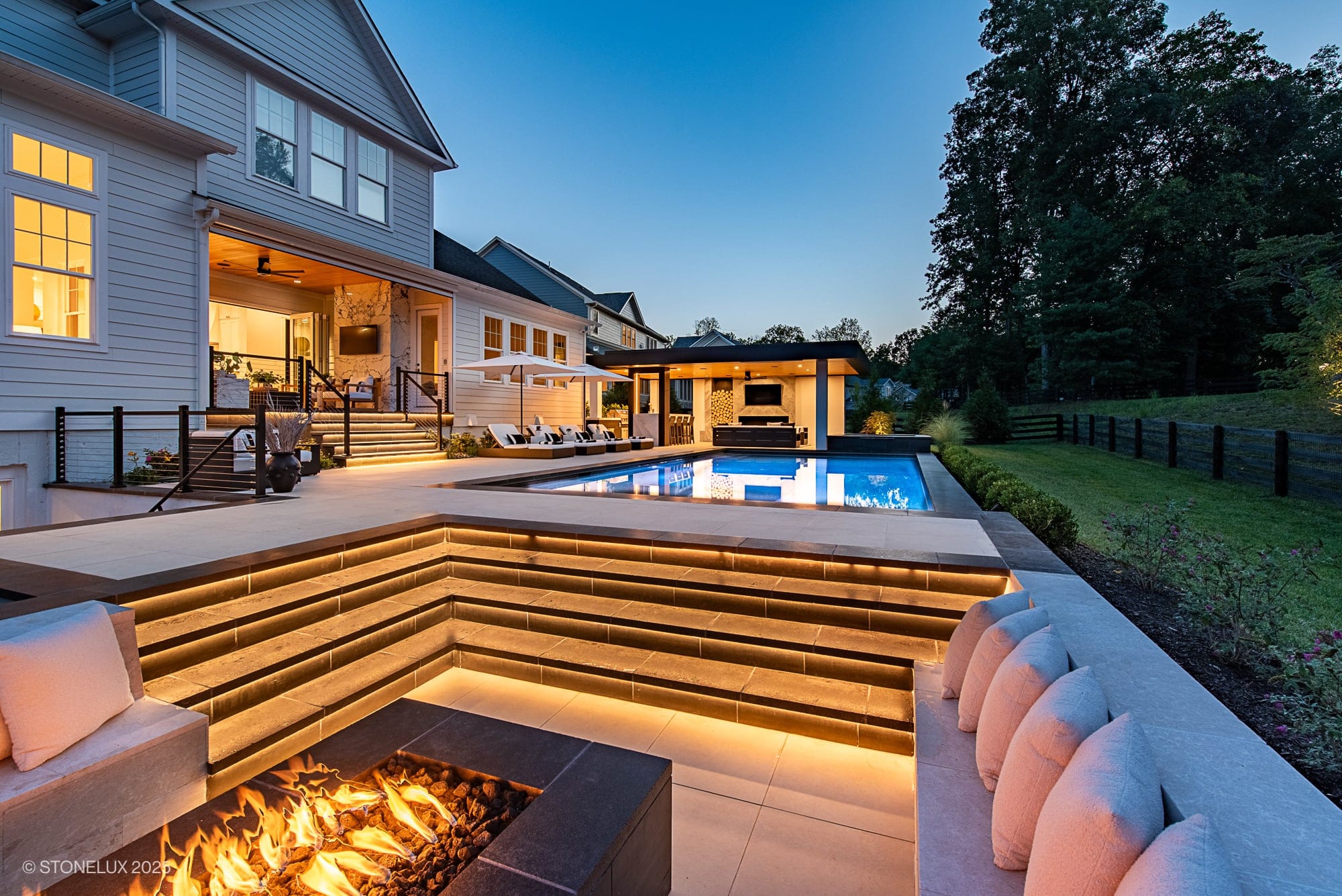 Aldie Modern Escape – Porcelain & Basalt Luxury Pool Pavilion: This modern backyard at dusk features a fire pit with built-in seating, illuminated steps, a swimming pool, lounge chairs, and a covered living area nestled among lush trees and landscaping.