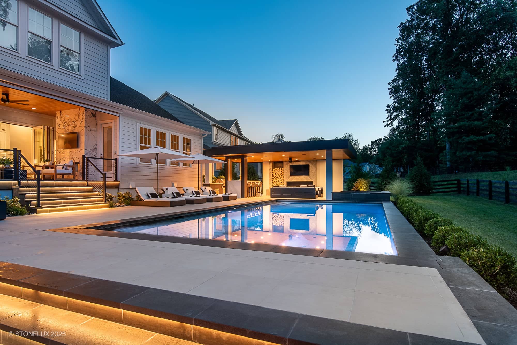 Modern backyard at sunset showcasing the Aldie Modern Escape – Porcelain & Basalt Luxury Pool Pavilion, with a swimming pool, poolside lounge chairs, covered outdoor seating, lush landscaped greenery, and warm lights illuminating the scene.