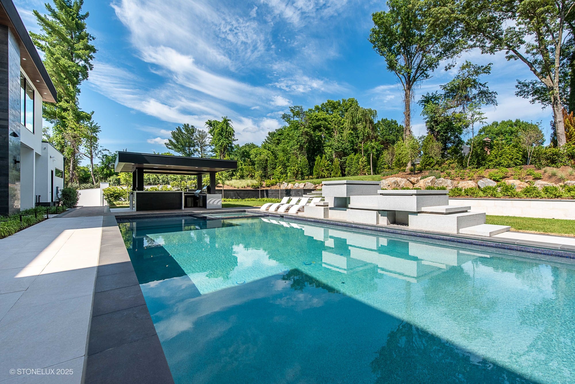 A modern outdoor swimming pool with clear blue water, surrounded by lounge chairs and shady seating areas, showcases Modern Hillside Elegance – McLean Retreat with Overland Pearl Porcelain & Black Basalt accents amid lush green trees.
