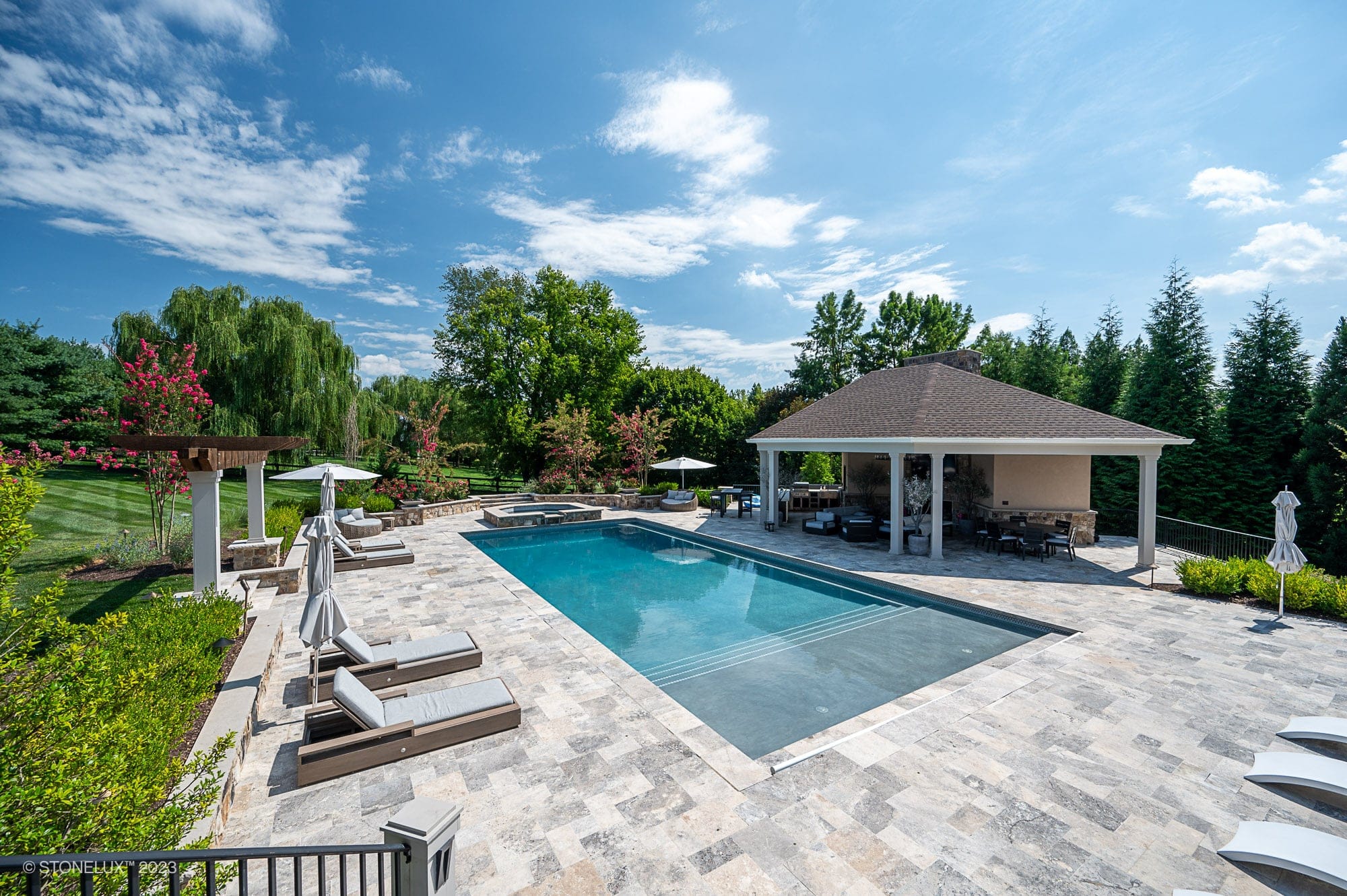 A luxurious backyard features a rectangular pool in the Silver Expanse: The Silver Travertine Pool Landscape, surrounded by stone tiles, lounge chairs, umbrellas, and a covered patio area with seating amid green trees under a blue sky.