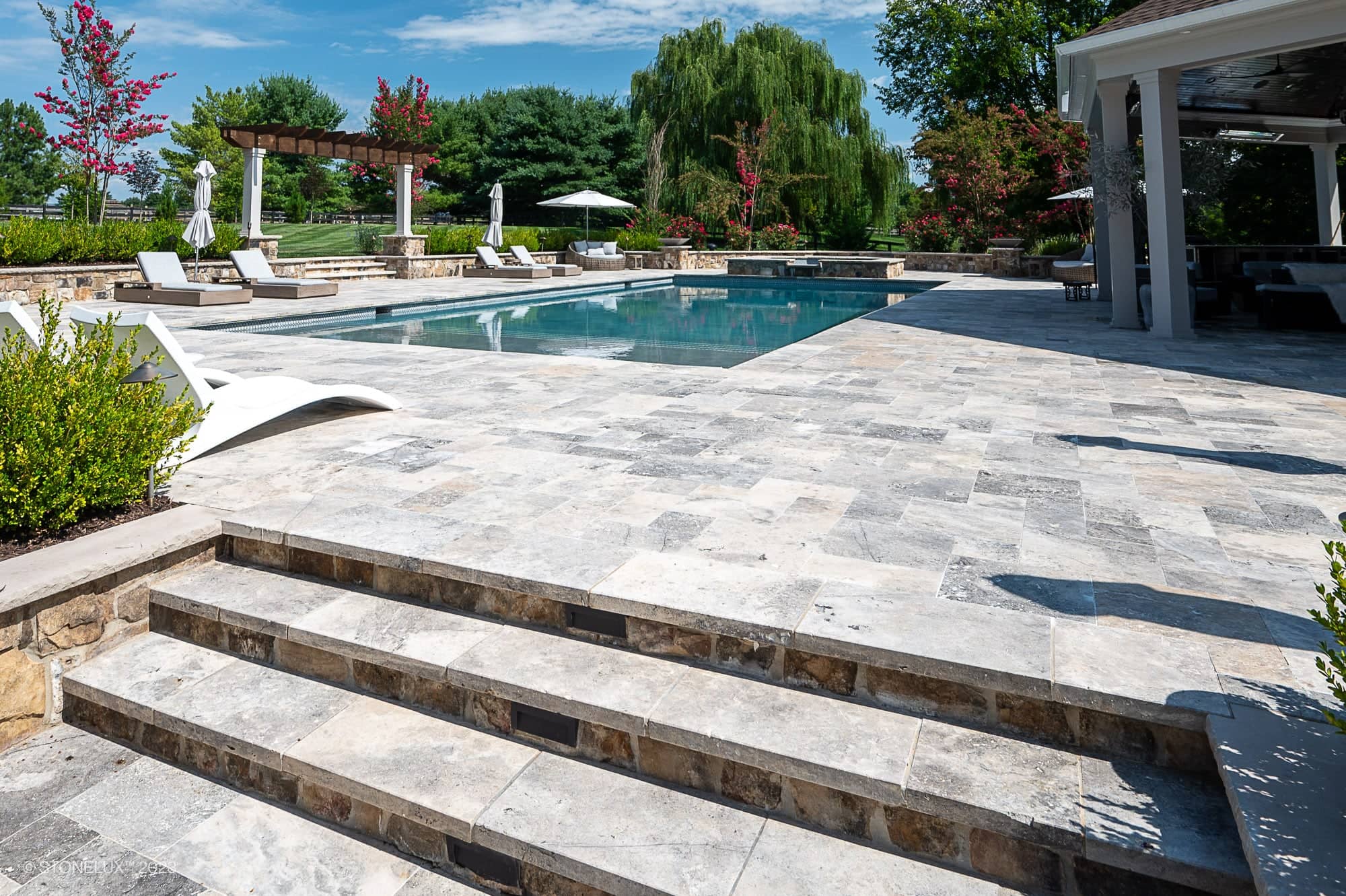 Outdoor swimming pool with lounge chairs, umbrellas, and landscaped greenery. The Silver Expanse: The Silver Travertine Pool Landscape features wide stone steps to the deck and a shaded seating area on the right under a roof.