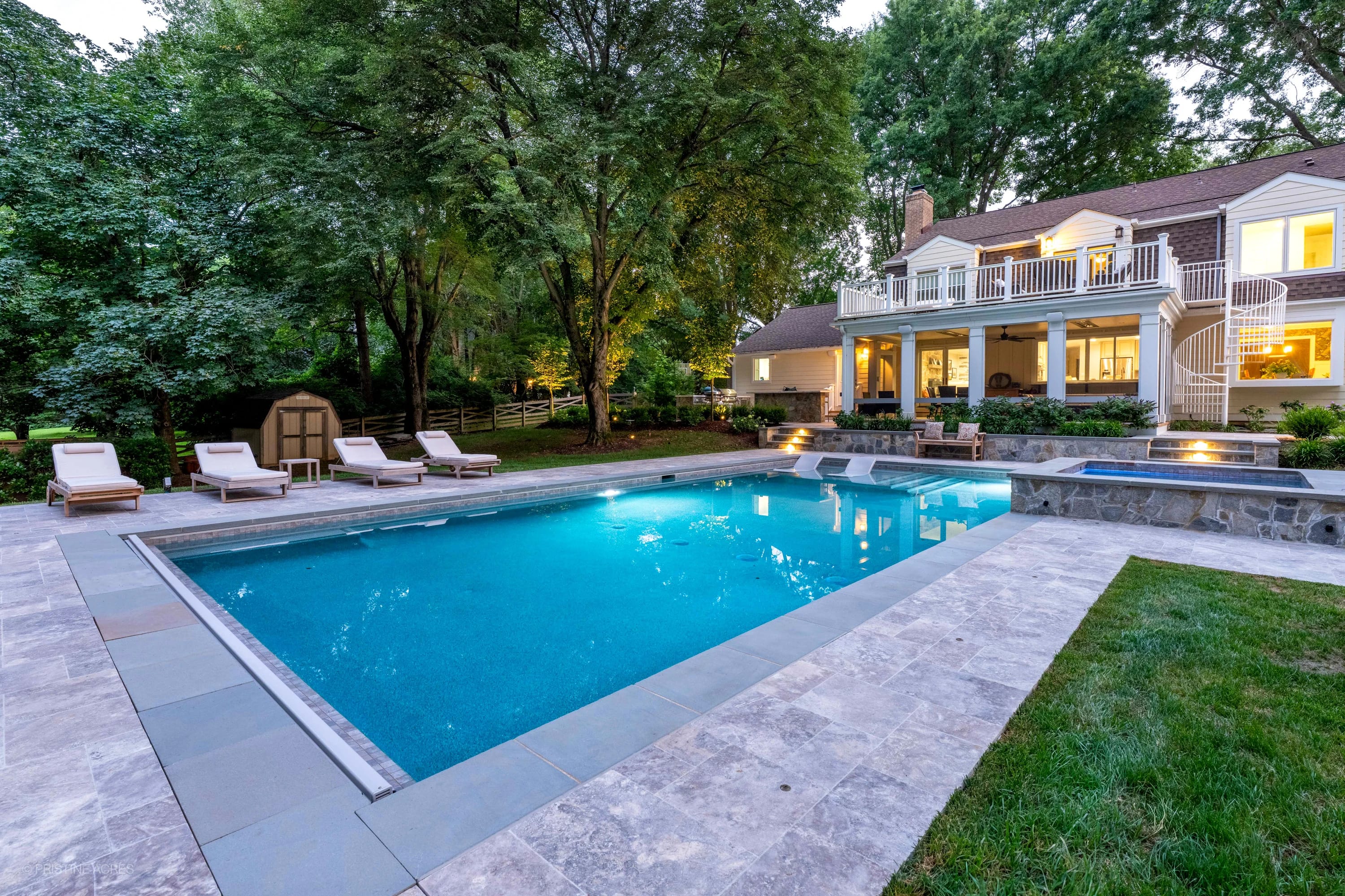 Vienna Pool Retreat featuring Silver Travertine and Bluestone combines elegant travertine pavers with bluestone accents around a serene blue pool. Set in a lush backyard with modern lounge chairs, mature trees, and a traditional house, creating a tranq...