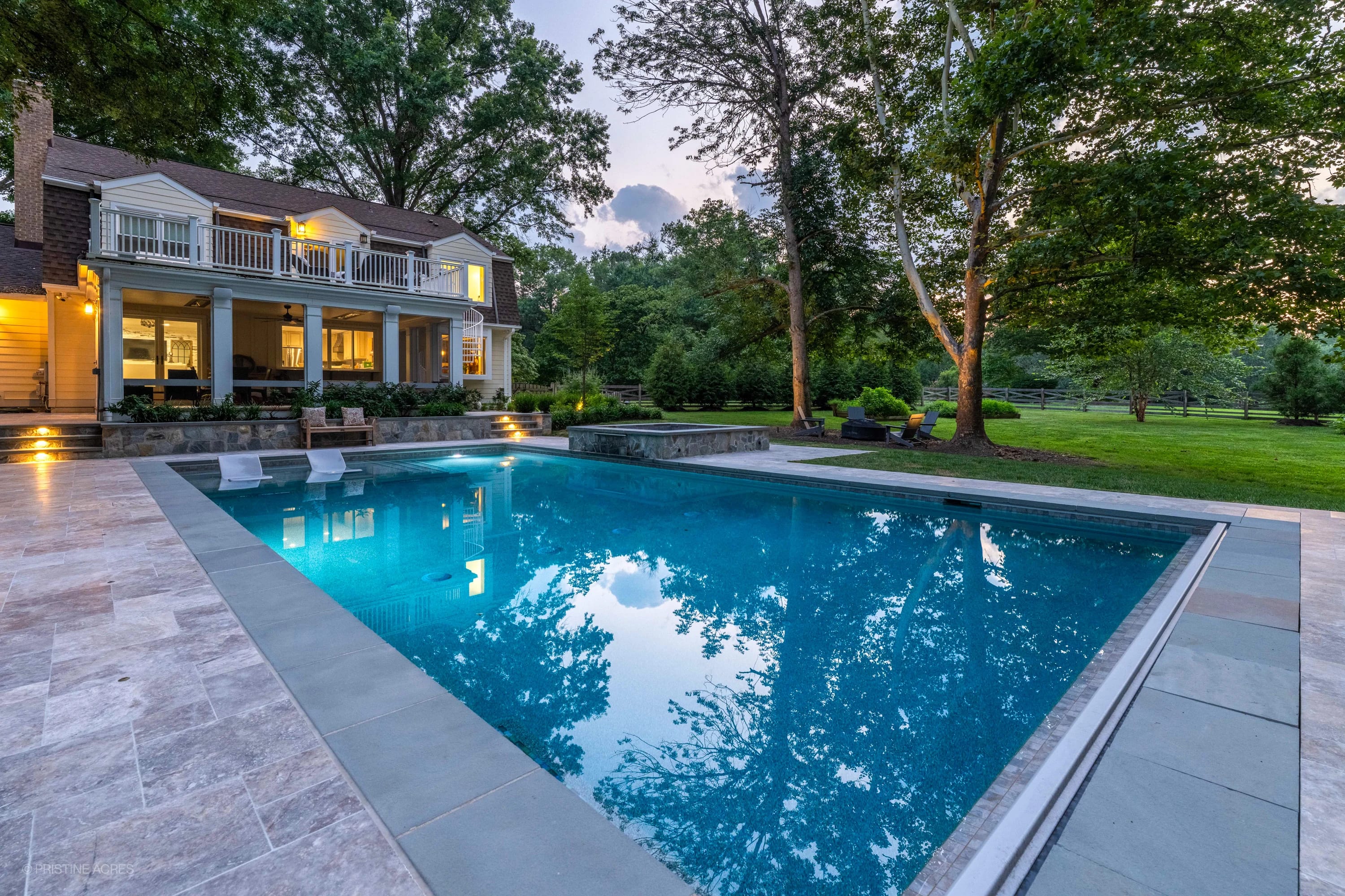 Luxurious Vienna Pool Retreat featuring Silver Travertine and Bluestone from Stonelux. The modern design includes elegant travertine pavers surrounding a vibrant blue pool, complemented by lush greenery and a traditional-style home, creating a serene b...