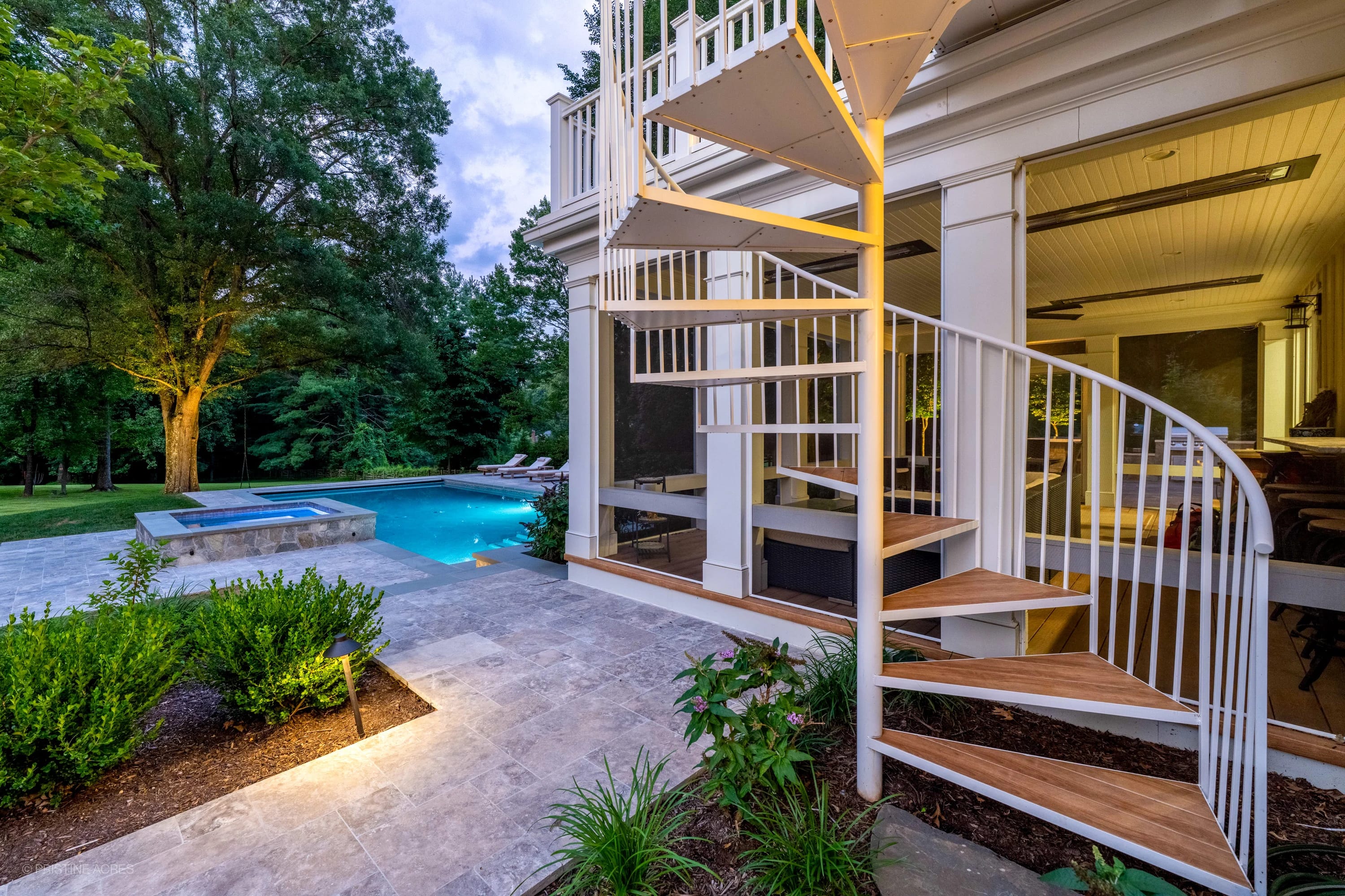 Vienna Pool Retreat in Vienna, VA features Silver Travertine pavers and bluestone, creating a modern outdoor oasis. The pool's blue water complements lush greenery and a spiral staircase, enhancing the serene backyard atmosphere.